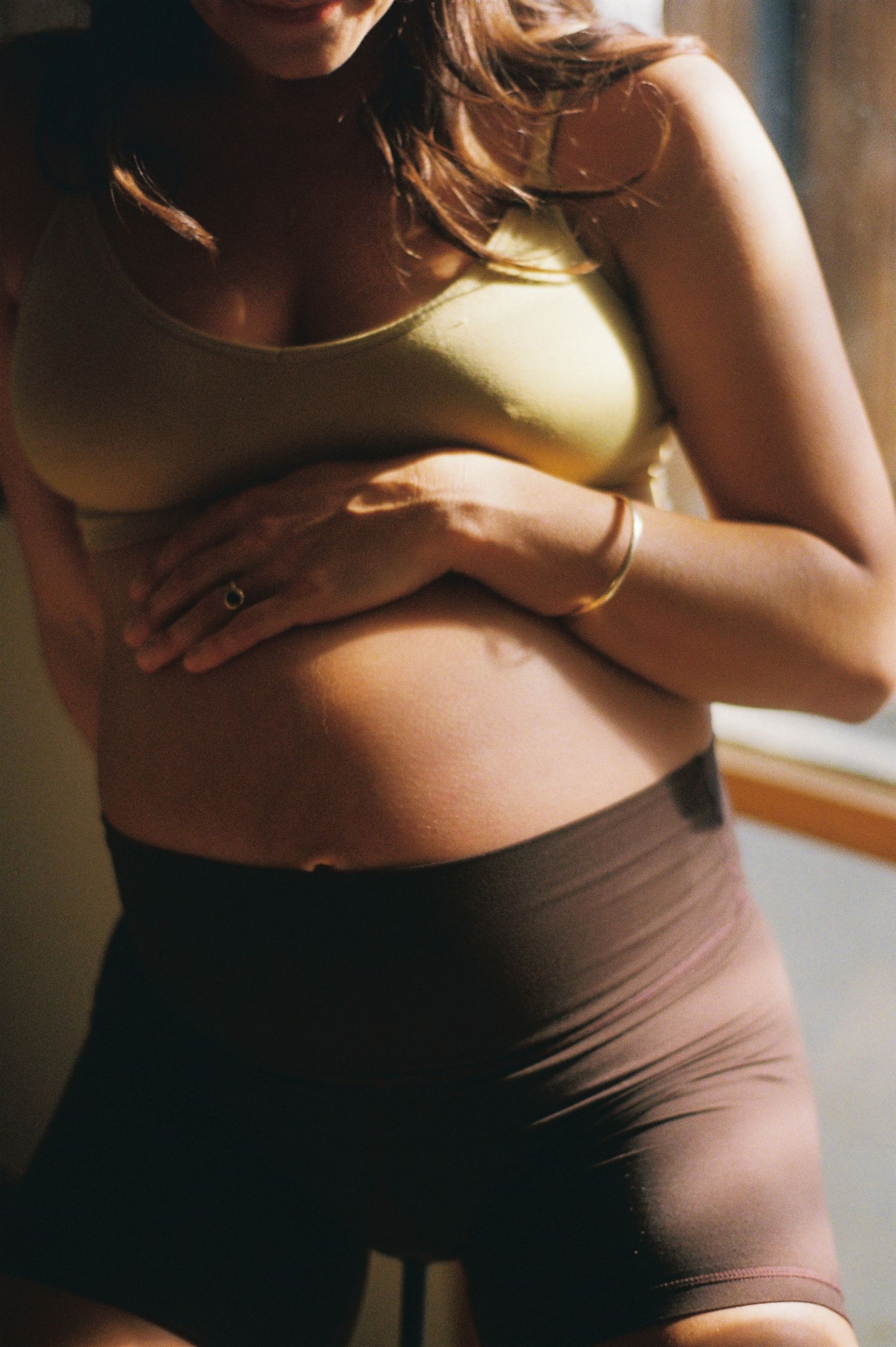 Pregnant woman in yellow sports bra and brown shorts cradling her baby bump, wearing gold jewelry.