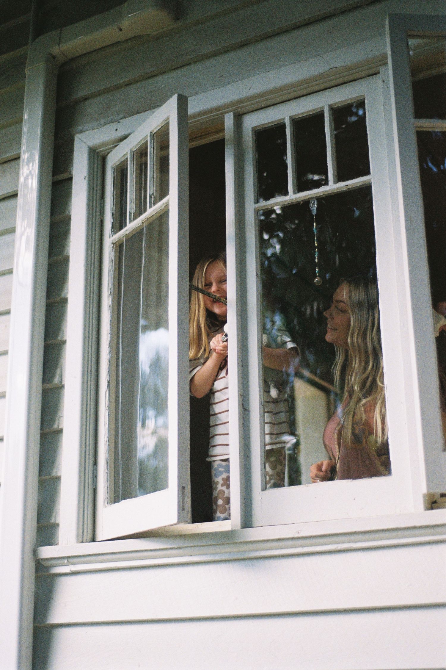 Two young women with long blonde hair laughing and chatting through an open white window of a wooden house.