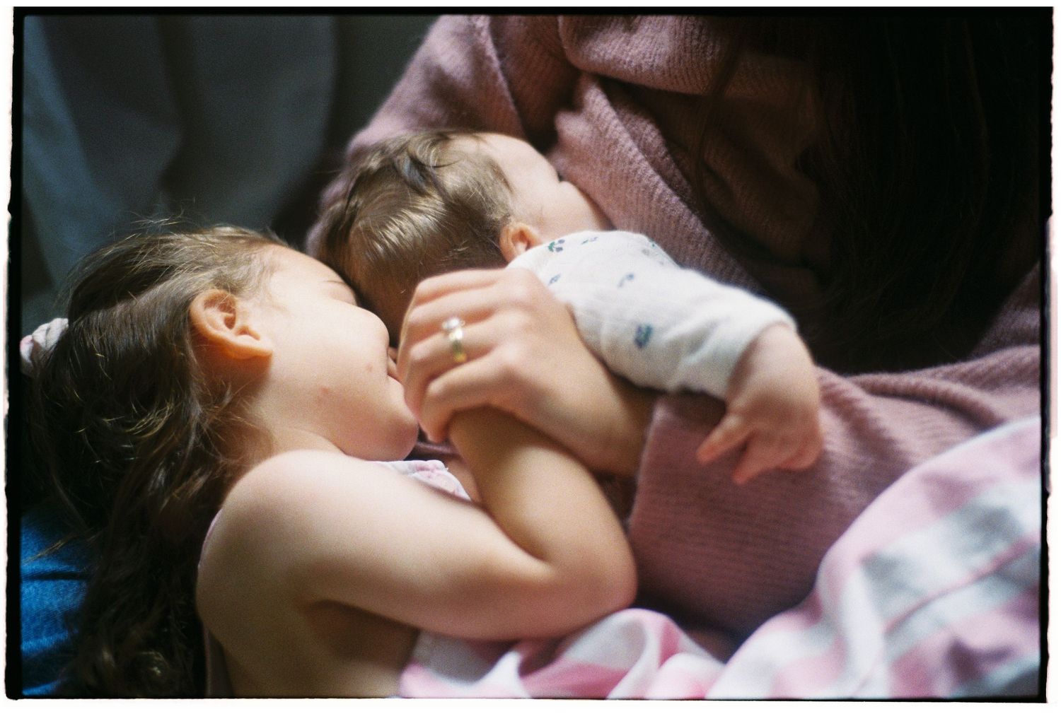 Young girl and baby sibling cuddling together in mother's arms on a pink blanket.