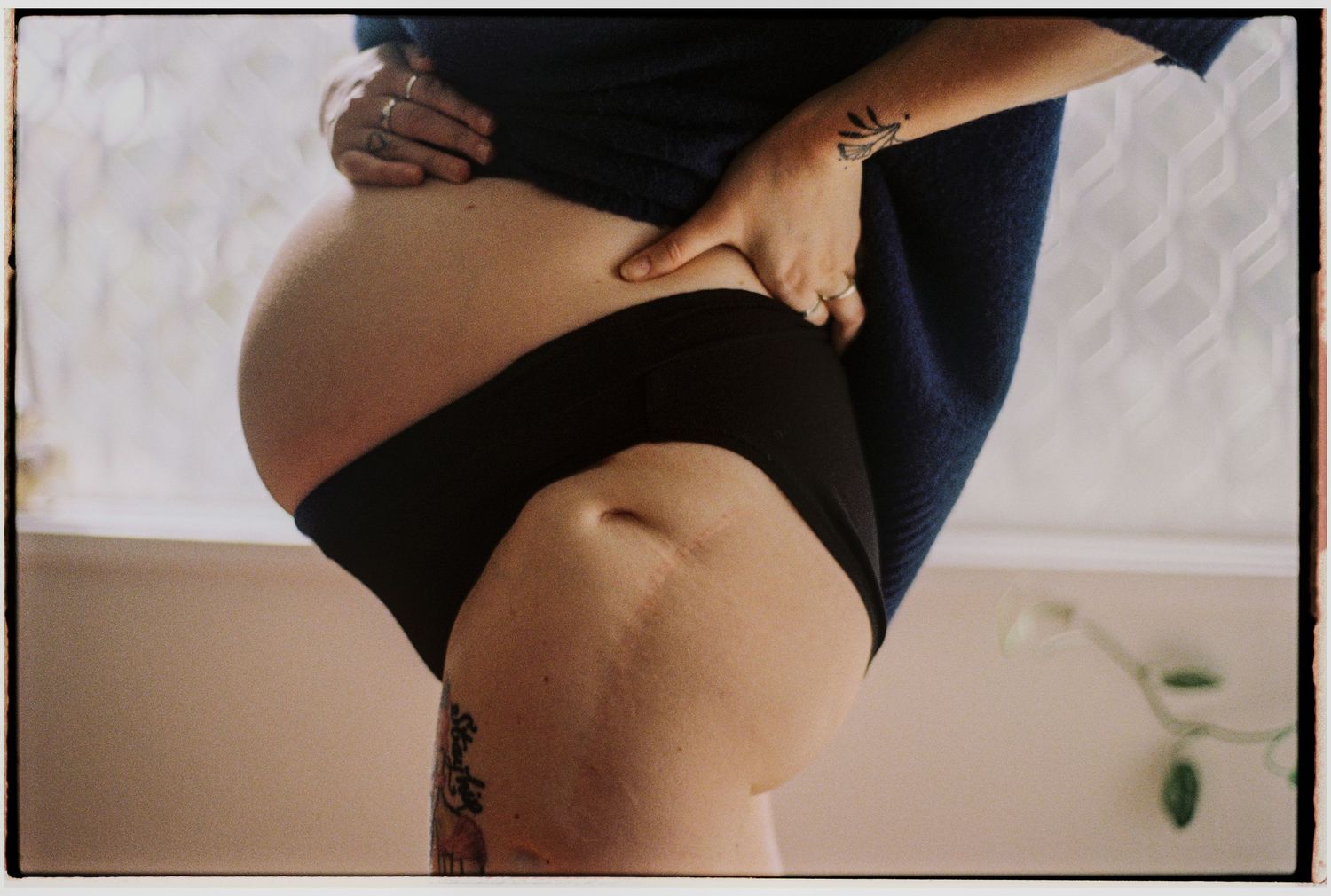 Pregnant woman in black underwear and blue top showing baby bump, with visible tattoos on leg and arm.