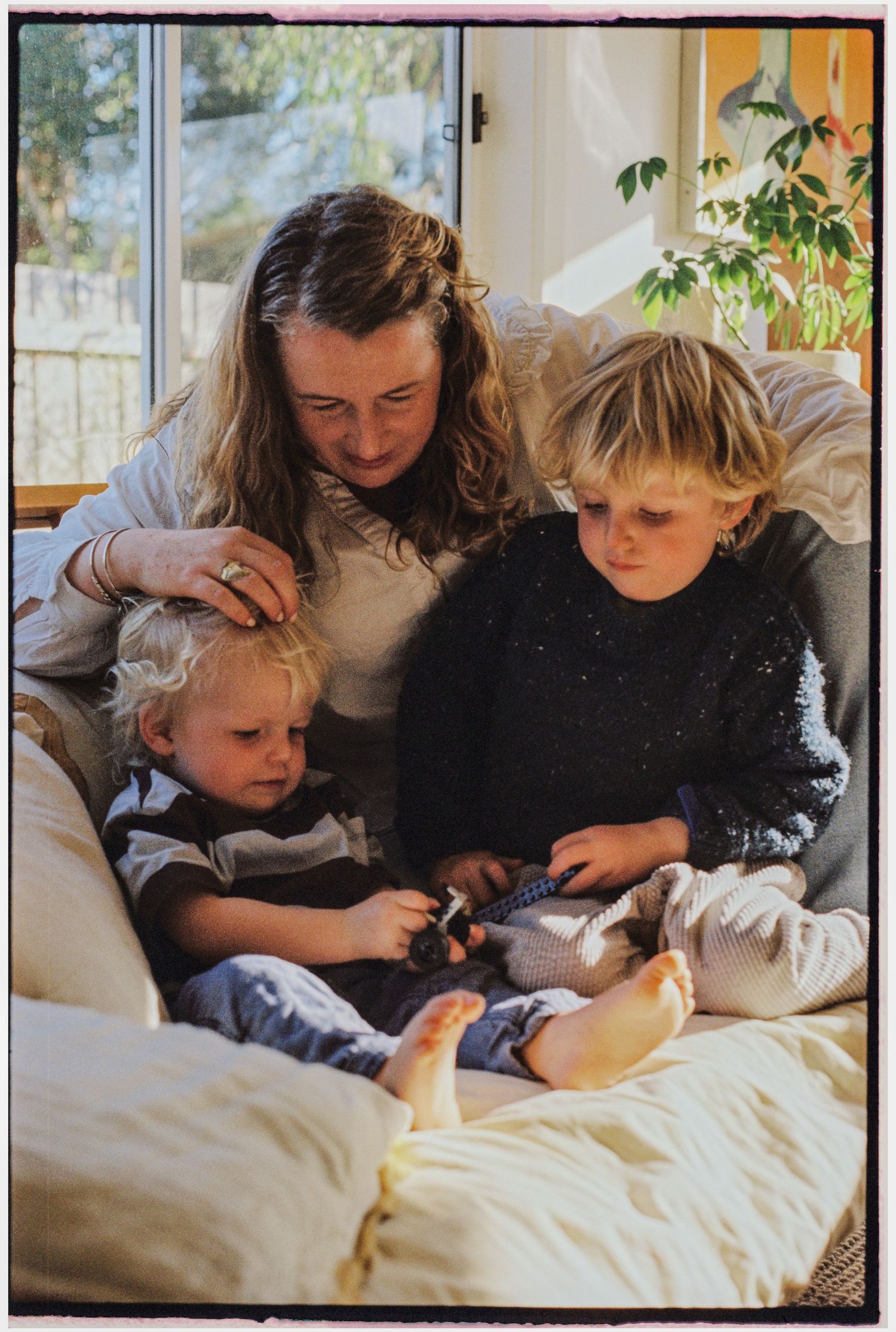 A mother sits on a couch with two young blonde children, looking at a small toy together near a sunny window.
