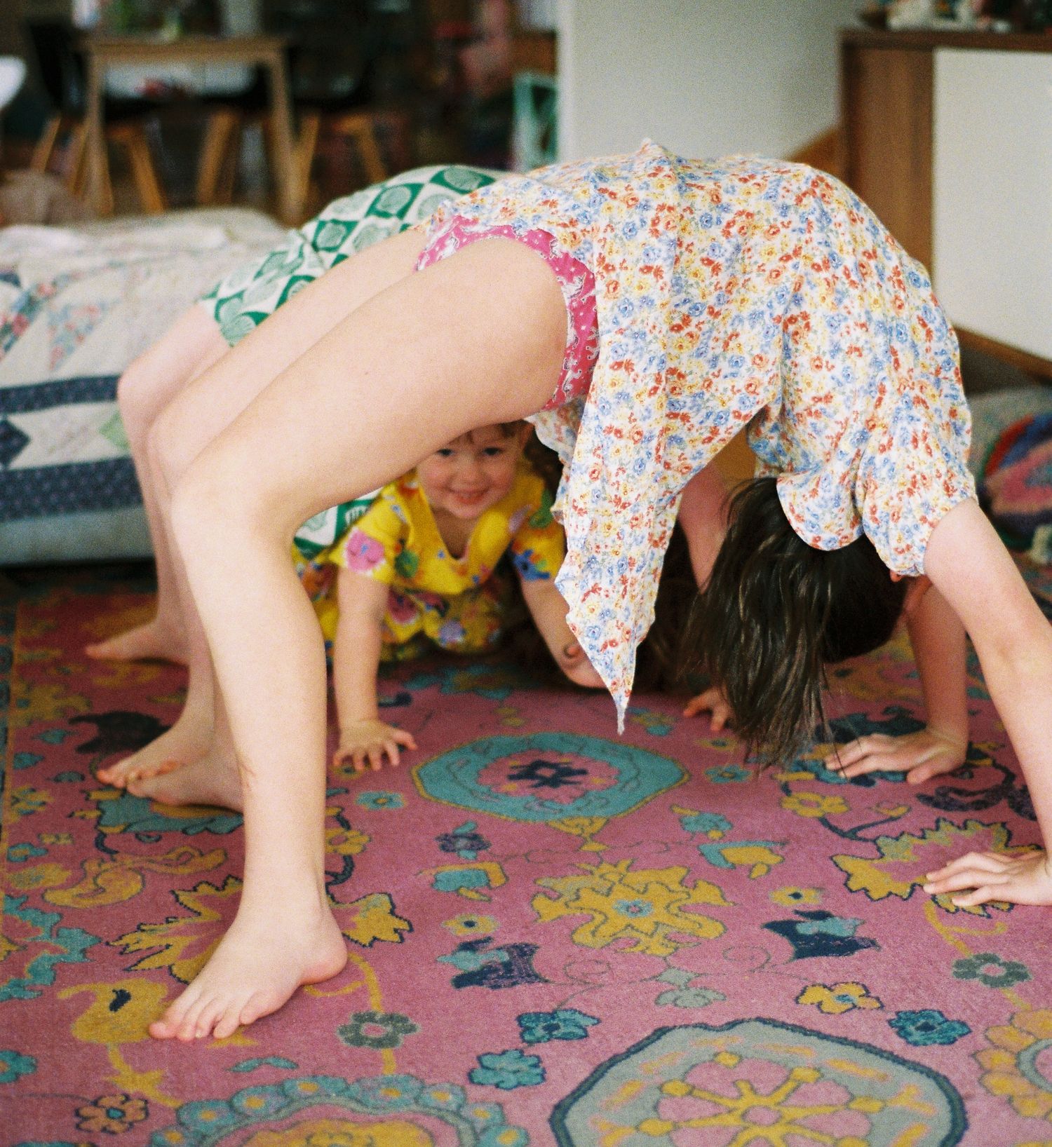 Child peeking through adult doing a backbend on a pink floral rug indoors.