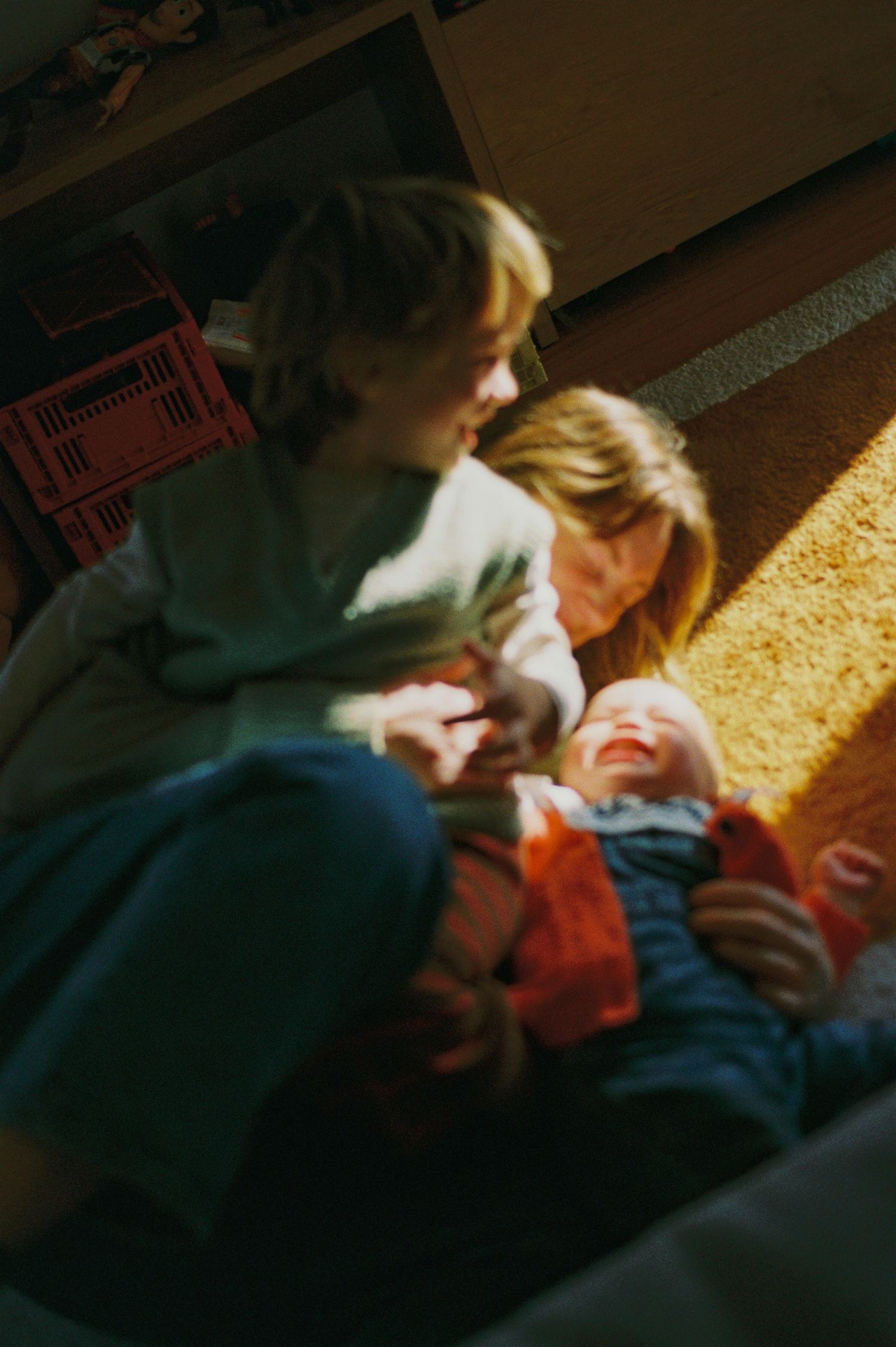 A mother and older child interact warmly with a laughing baby on a sunlit carpet floor.