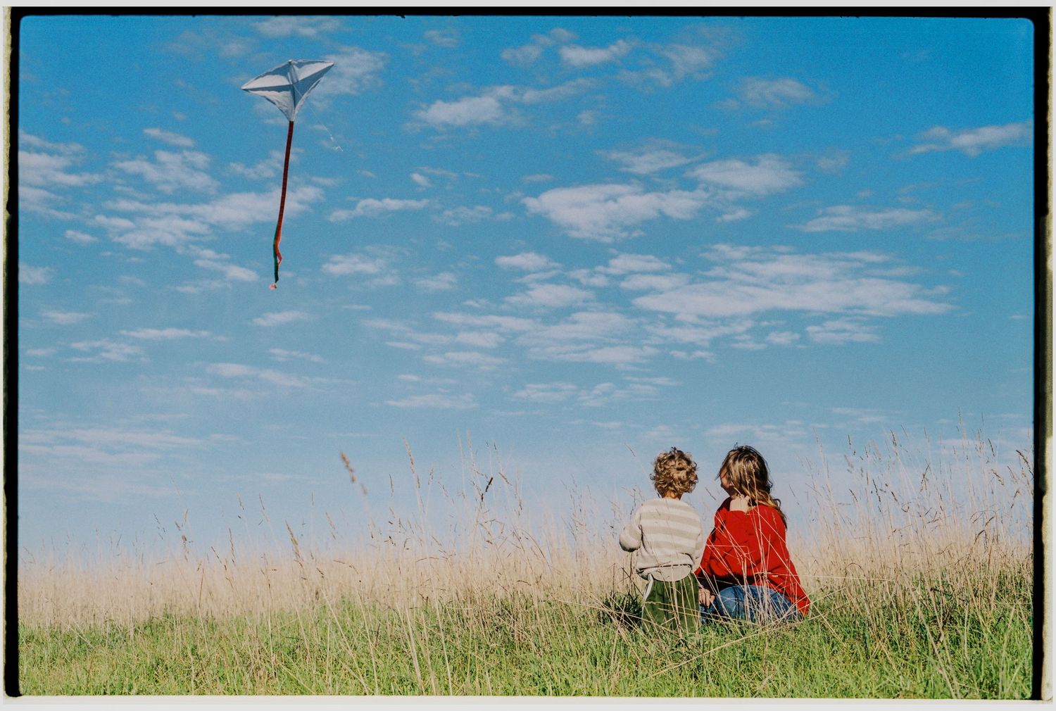 Two children sit in a grassy meadow flying a white kite against a bright blue sky with wispy clouds.
