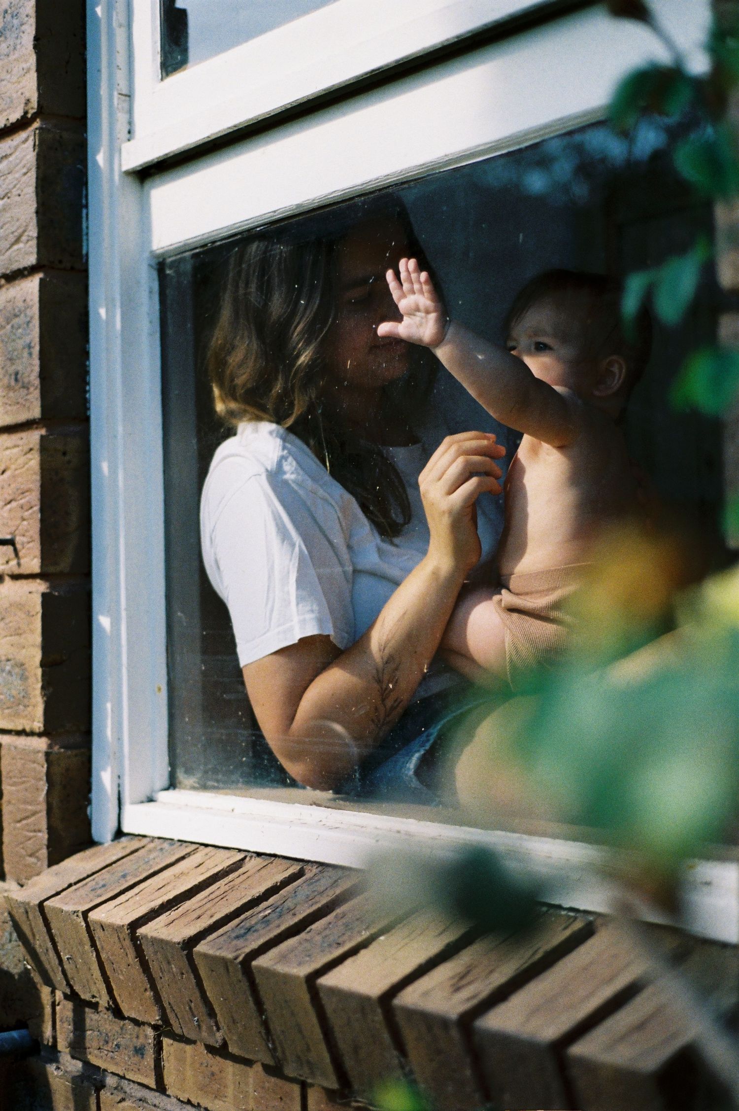 A parent holds a baby near a sunlit window, with the child's hand pressed against the glass.