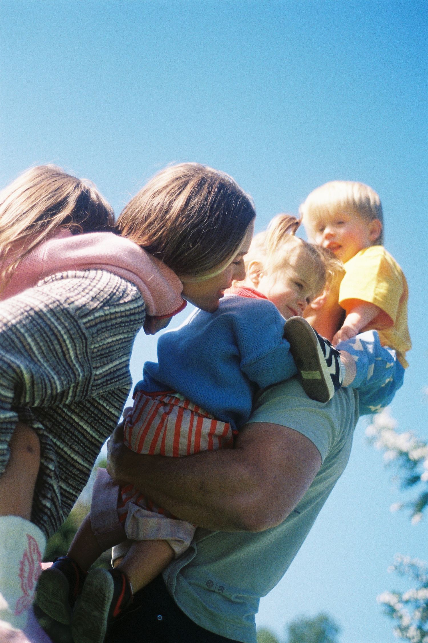 A parent holds two young blonde toddlers outdoors under a clear blue sky on a sunny day.