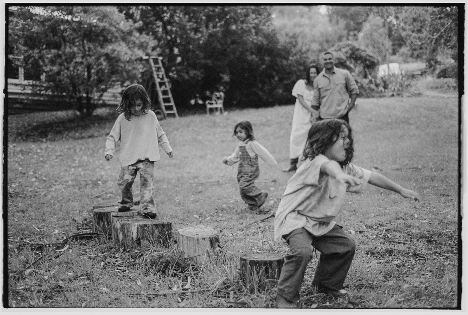Children playing on tree stumps in a grassy yard while adults watch in background, black and white vintage photo.
