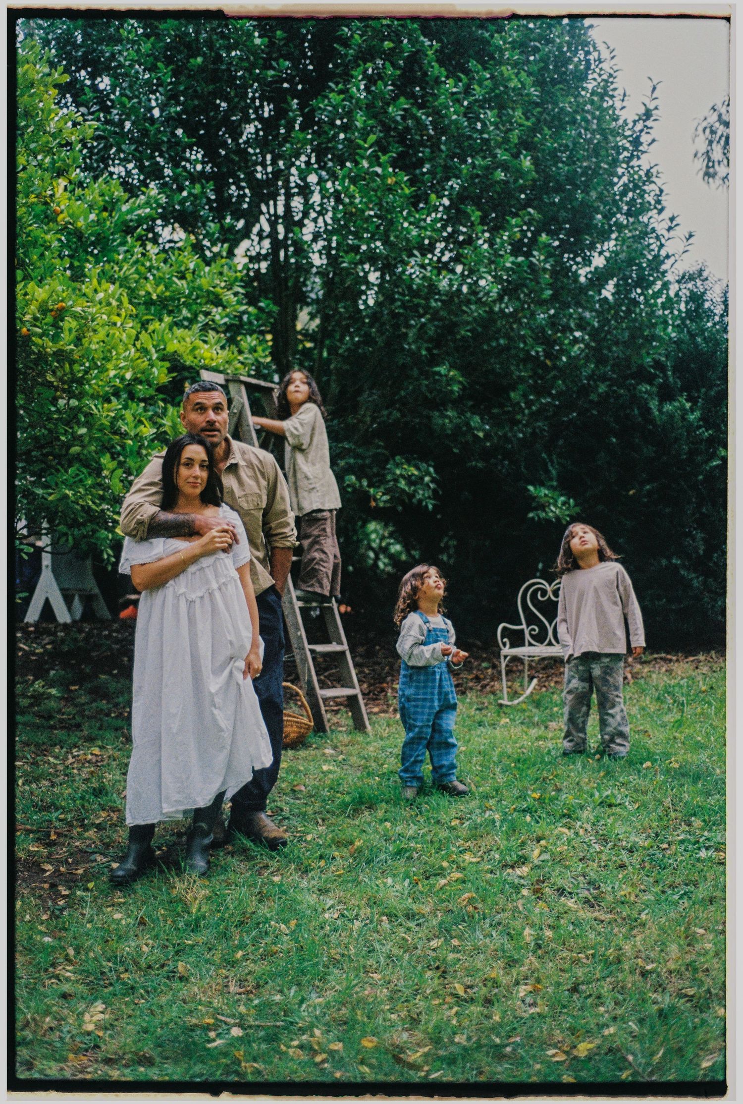 Family of five poses in lush garden with ladder, children playing near ornate chair and tall trees.
