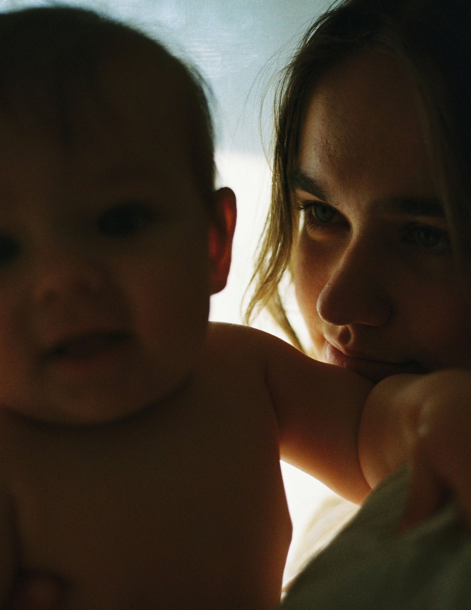 A young woman tenderly holds a baby close, both bathed in warm, dramatic side lighting in an intimate portrait.