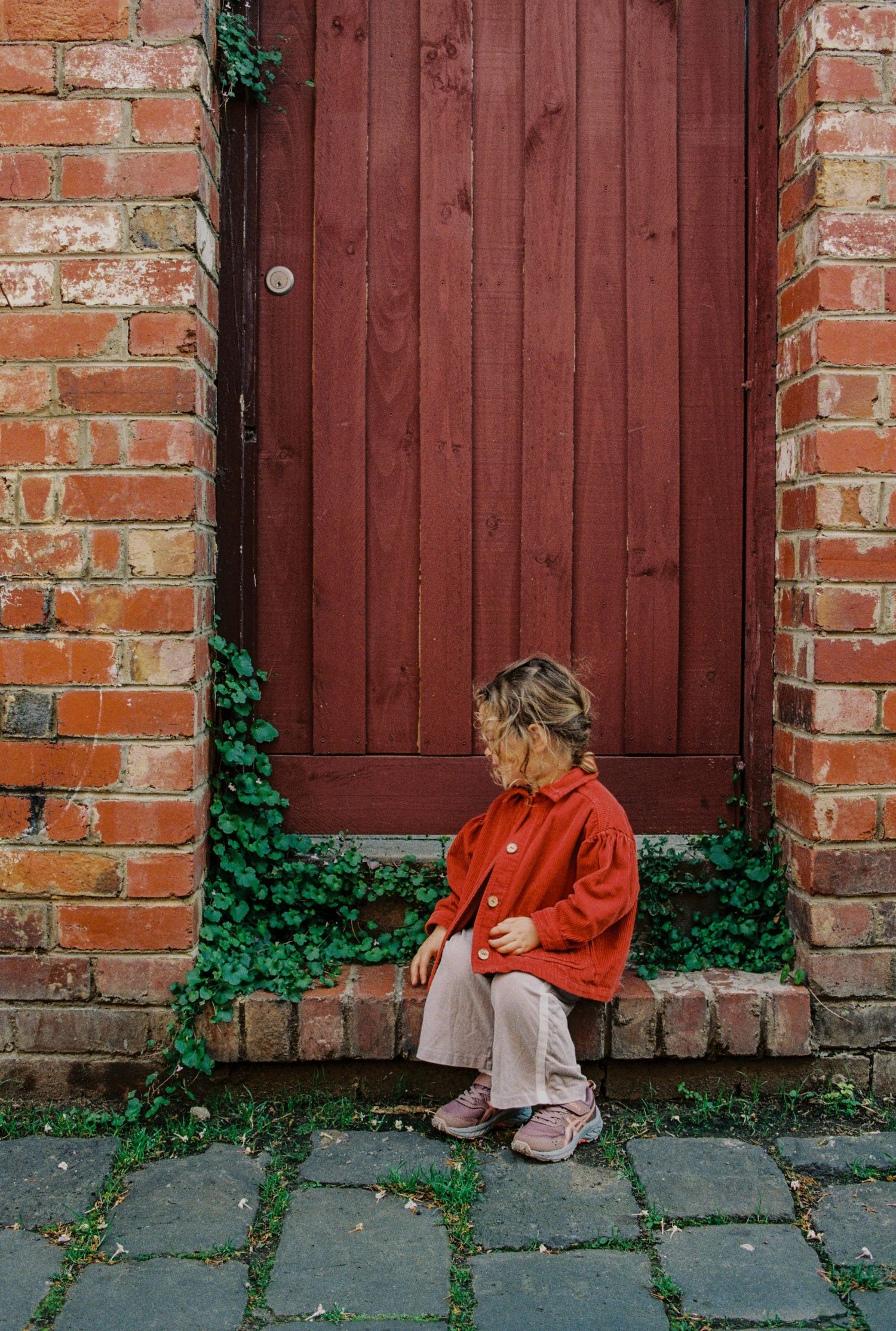 A young child in a red jacket sits on a brick doorstep beside a dark red wooden door with green ivy.