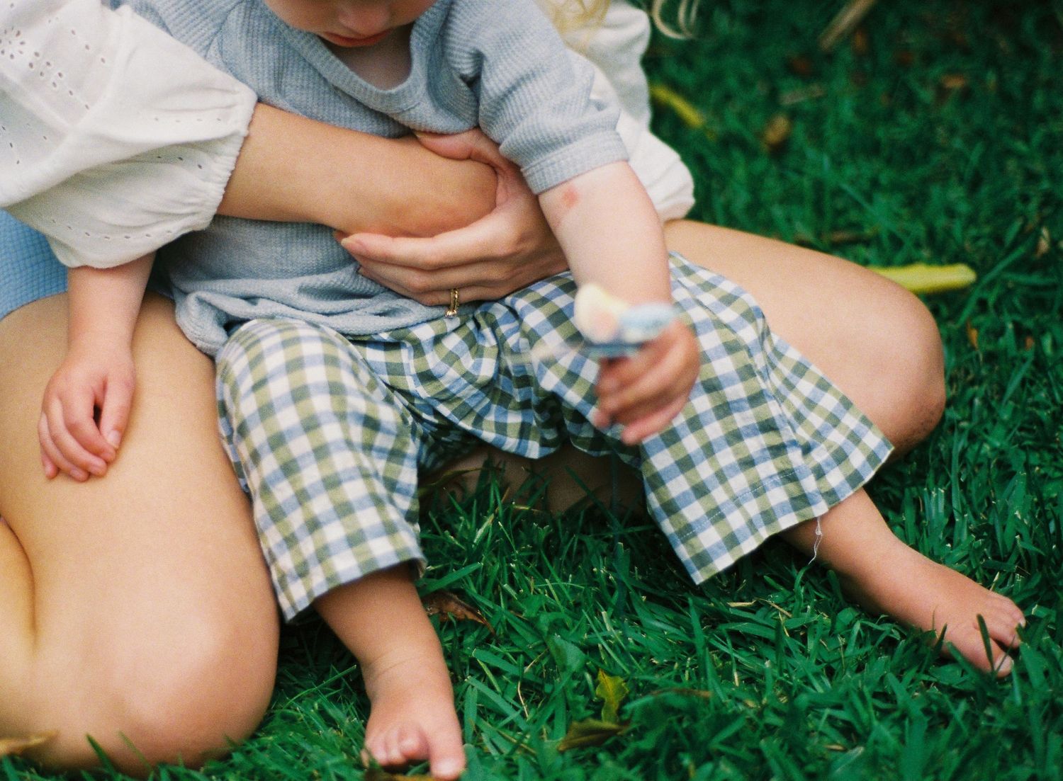Mother holding toddler in blue gingham pants sitting on lush green grass outdoors.