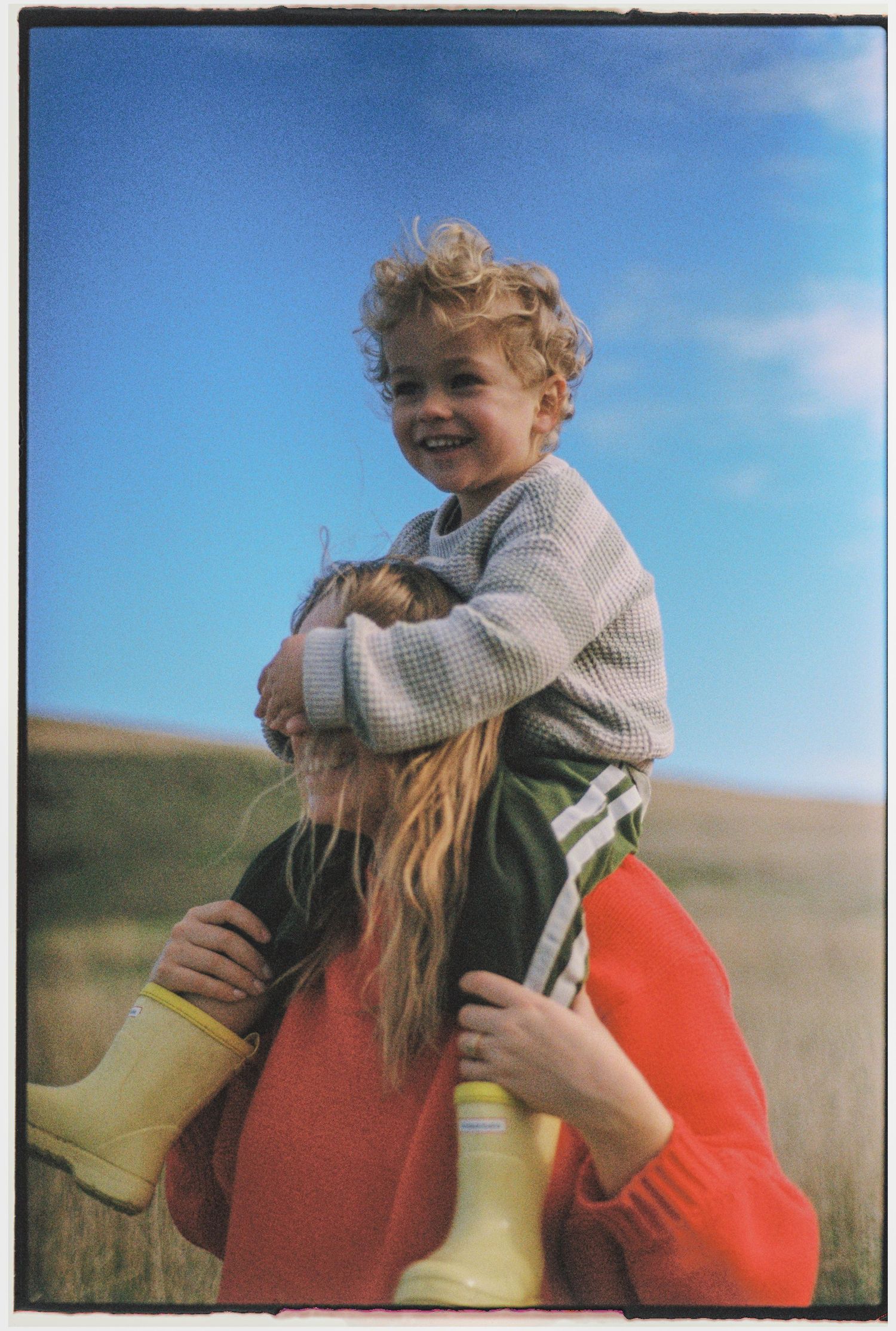 A smiling child sits on an adult's shoulders in a sunny field, wearing a sweater and green pants with yellow boots.