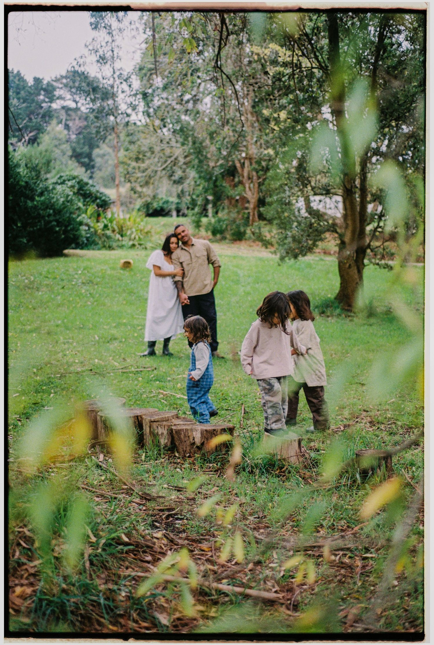Family with two children playing on tree stumps in a lush green garden park setting.