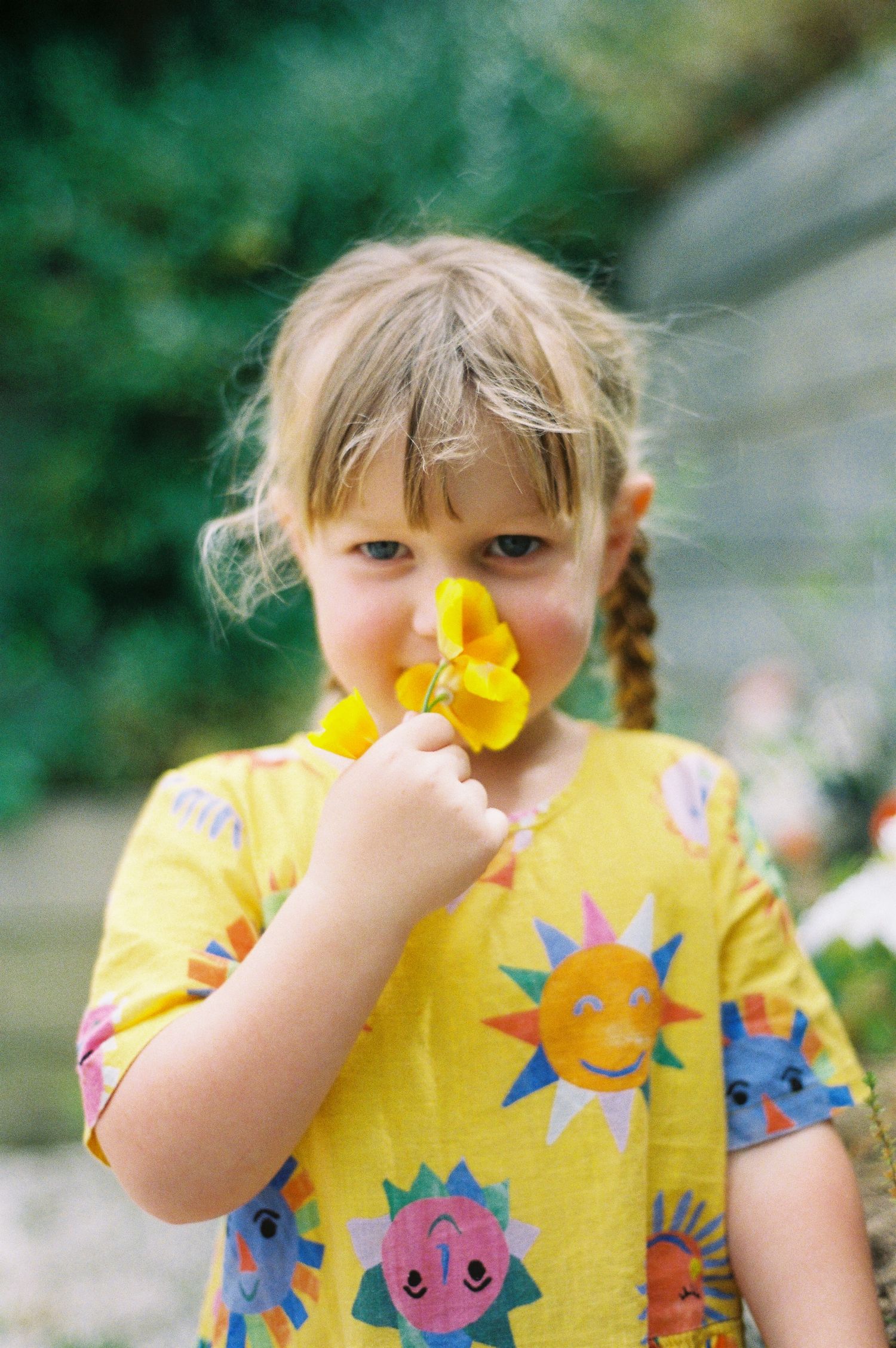 Young girl with braids in yellow sun-print shirt smelling a bright yellow flower outdoors.