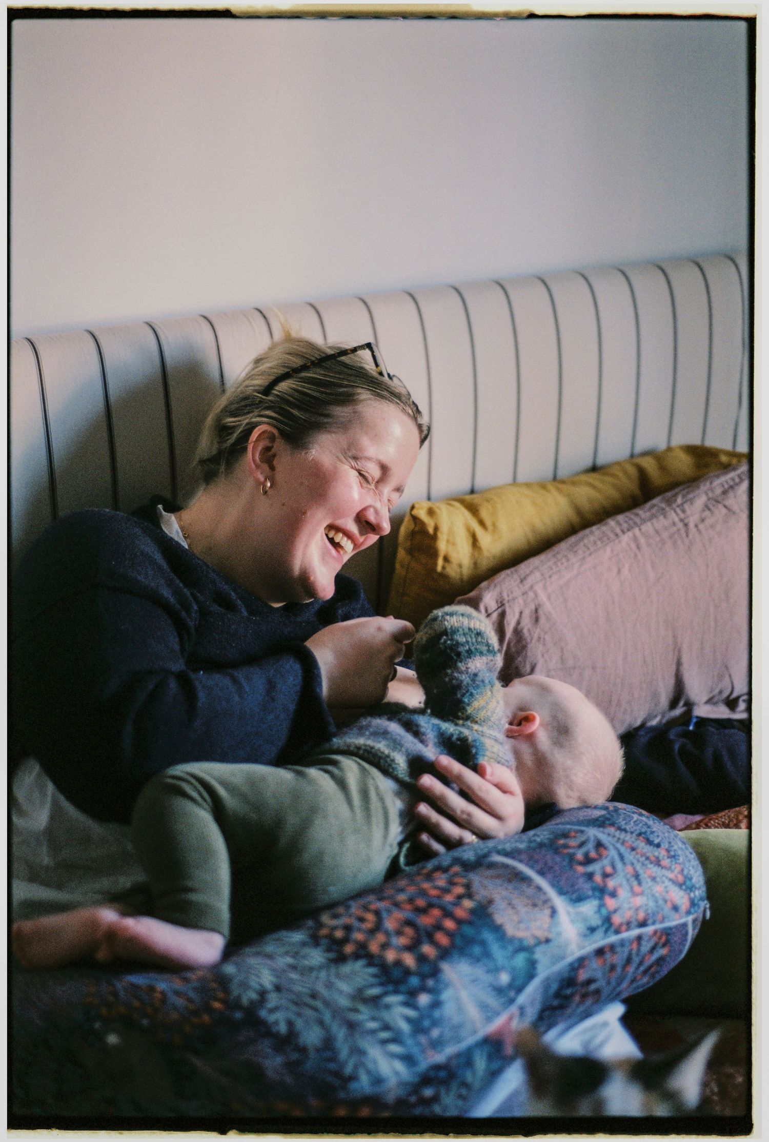 A laughing woman cuddles and plays with a baby on a cozy bed with colorful pillows.