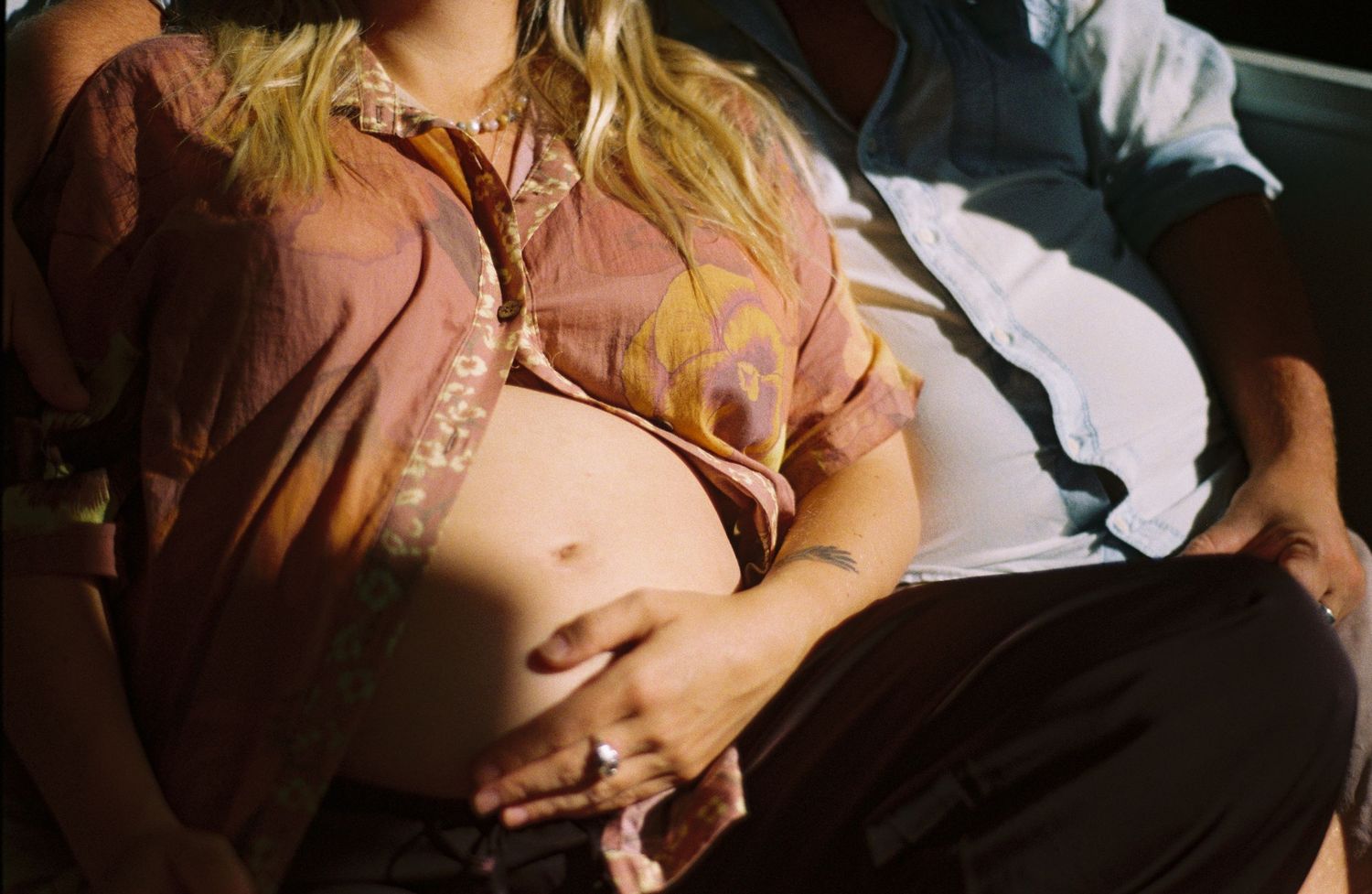 Pregnant woman in pink floral shirt cradling baby bump, sitting beside partner in warm sunlight.