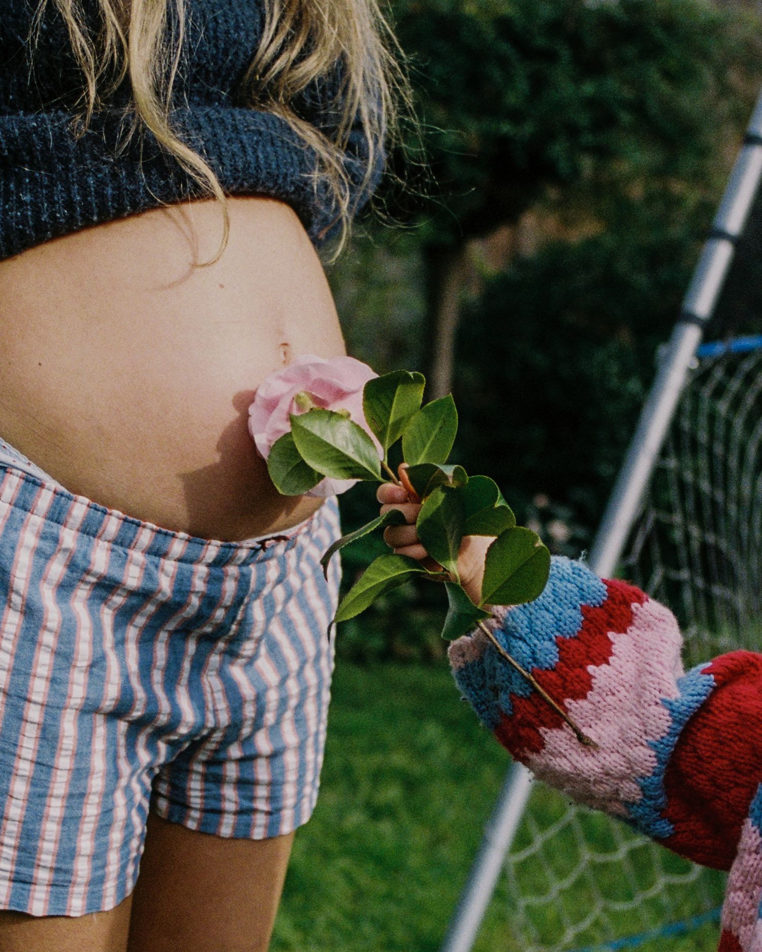 Pregnant woman in striped shorts outdoors as a child offers her a flower sprig while wearing colorful knit mittens.