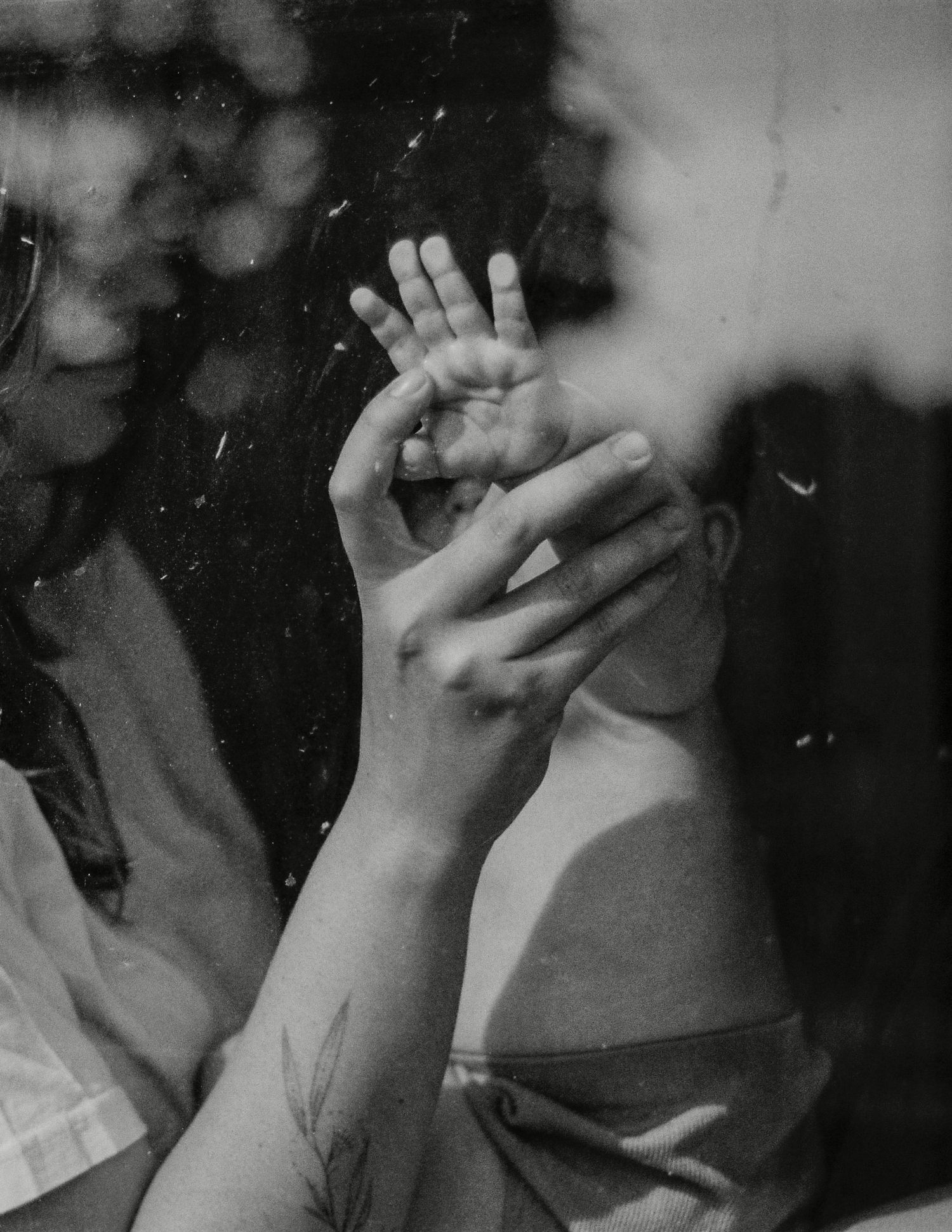 Black and white photo of two hands touching through a rain-speckled window, with a floral tattoo visible.