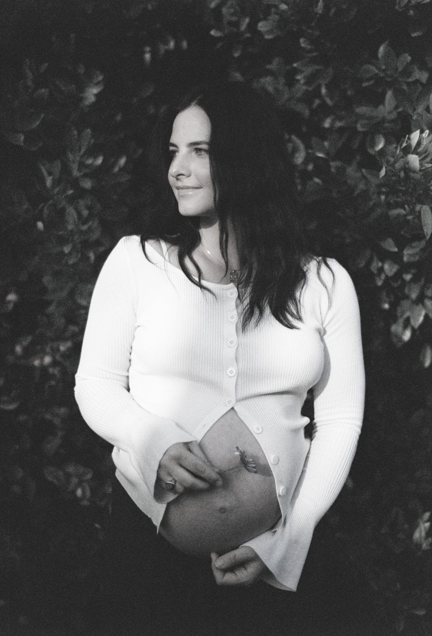 Black and white maternity photo of a woman in white cardigan cradling her baby bump outdoors.