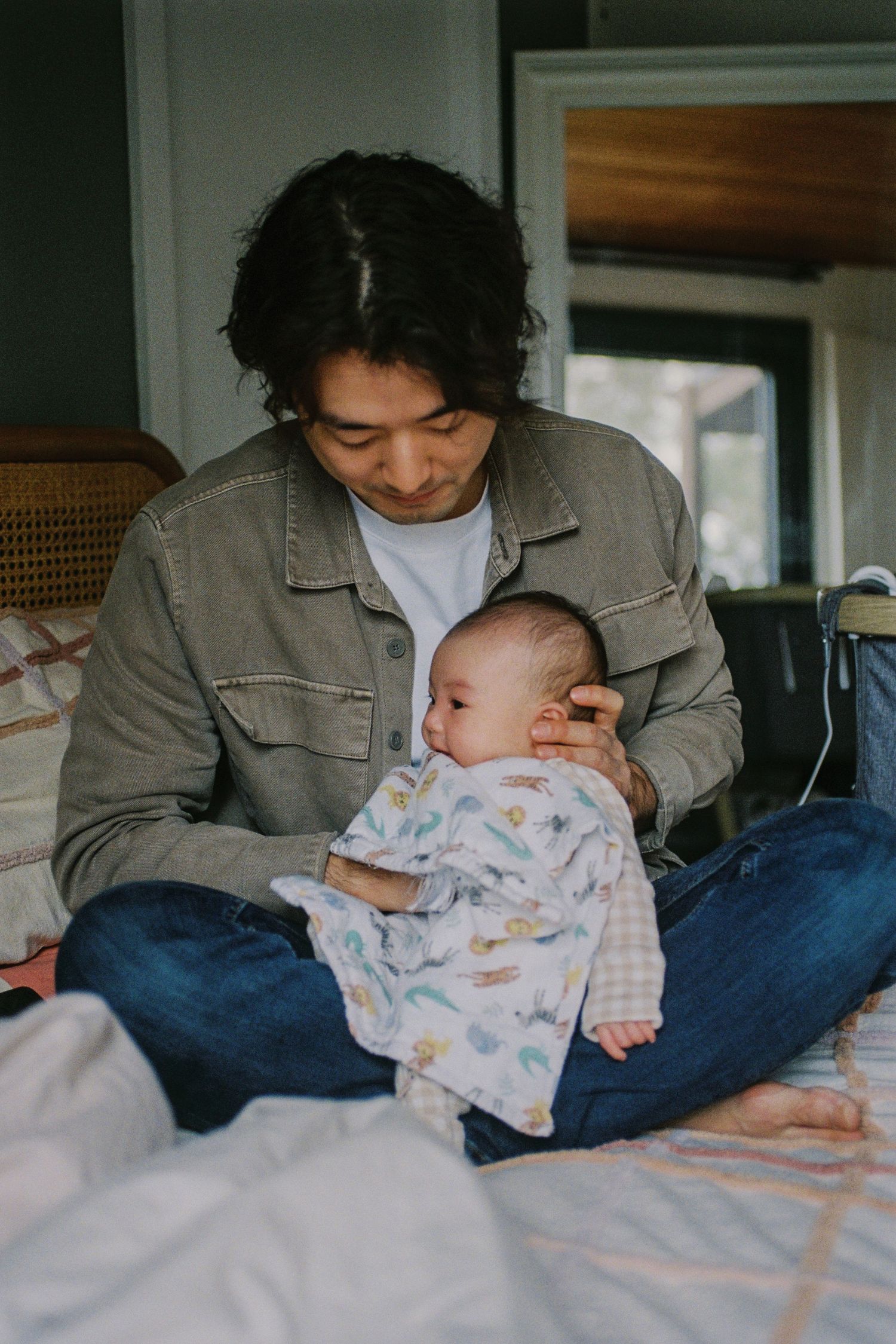 A young man with dark hair sits cross-legged on a bed, tenderly cradling a newborn baby wrapped in a floral blanket.