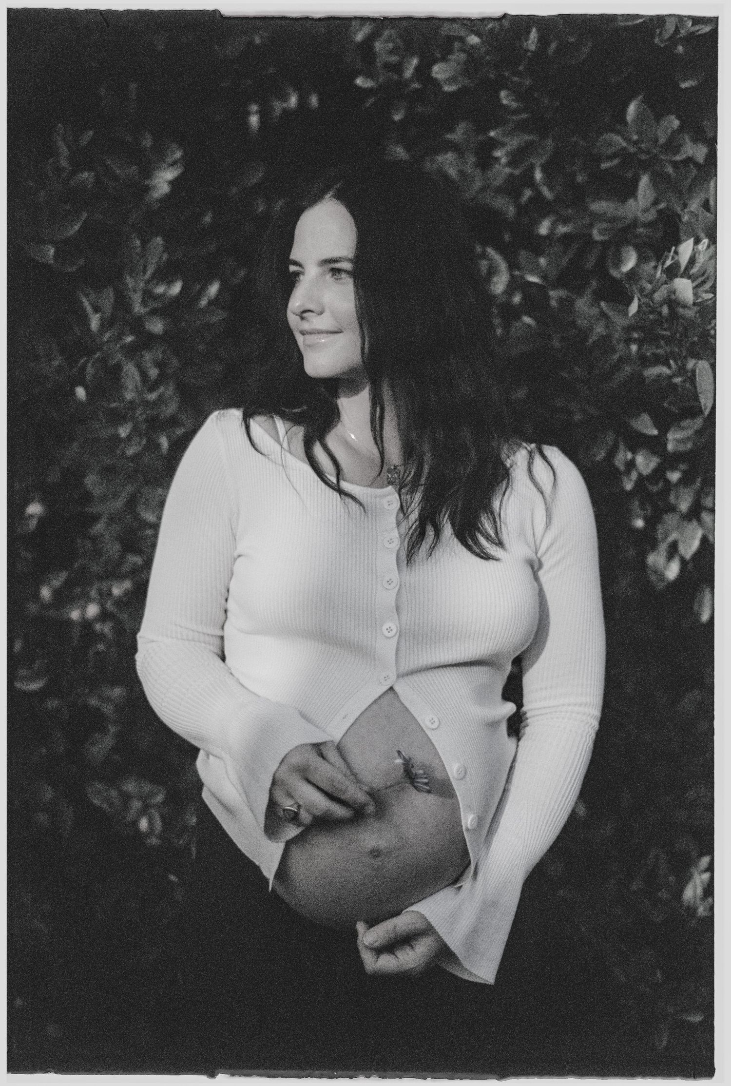 Black and white photo of a pregnant woman in a white cardigan cradling her baby bump, standing outdoors near leafy bushes.