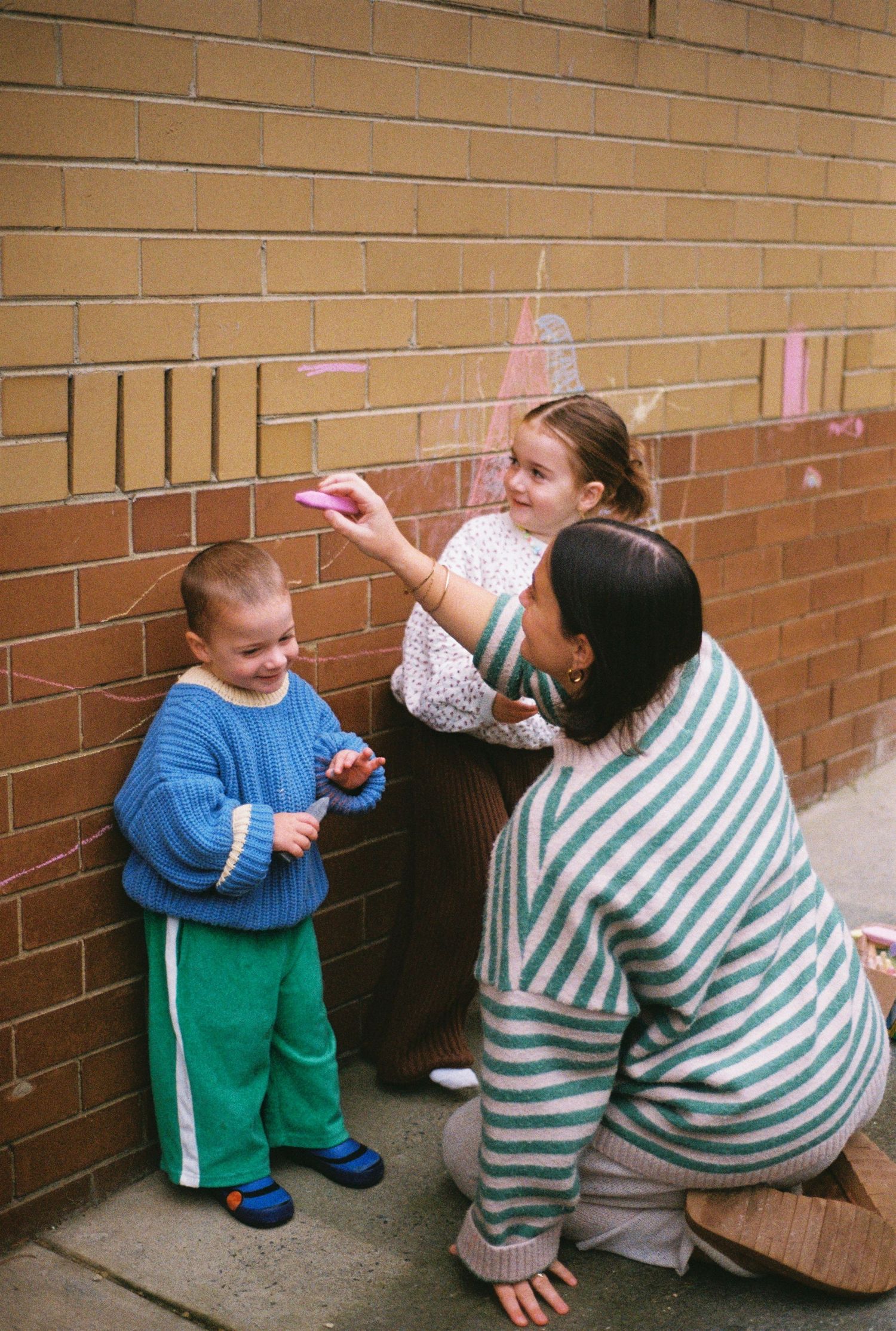 A woman crouches beside two children drawing with chalk on a brick wall outdoors.