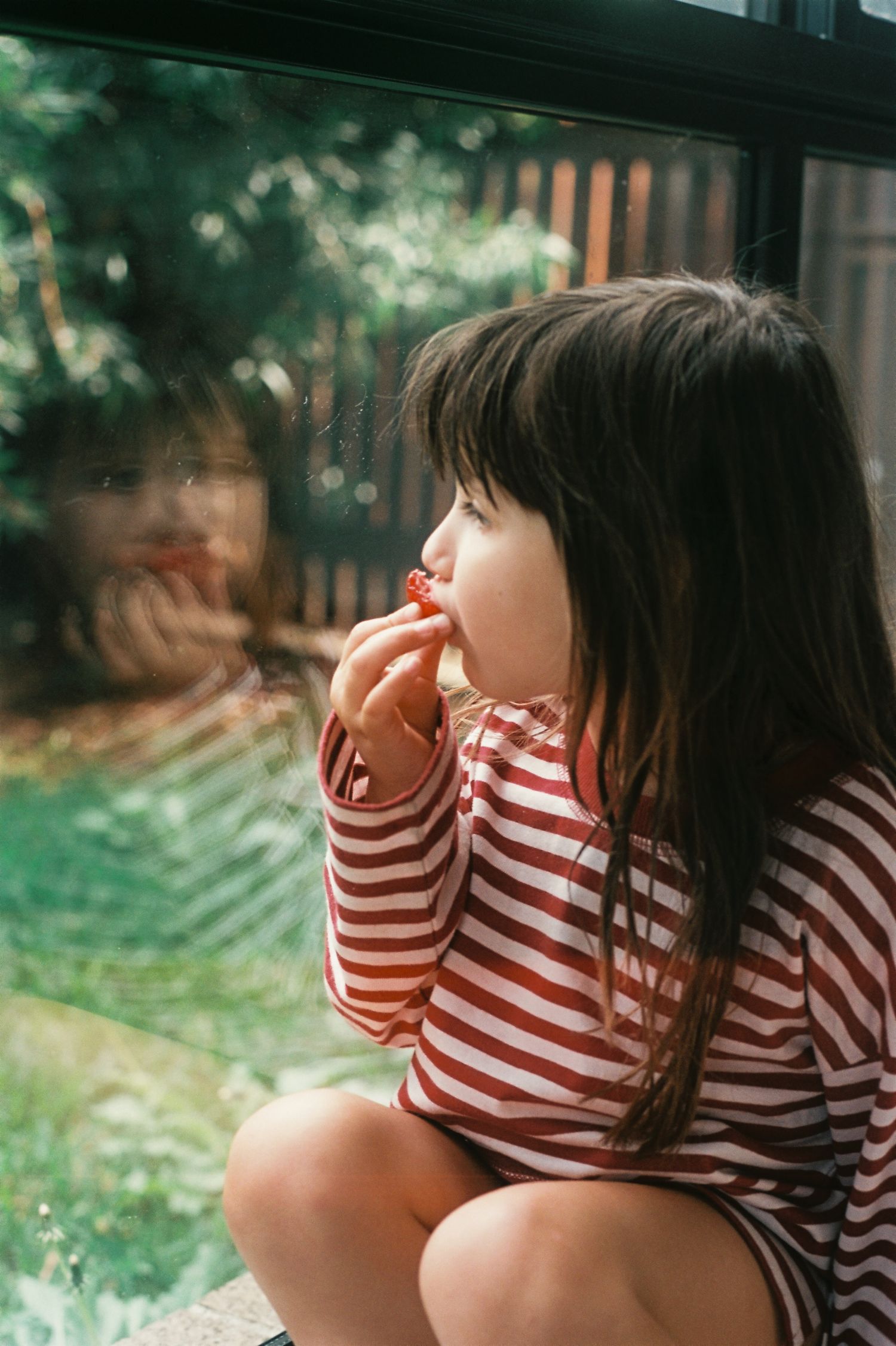 Young girl in red striped shirt eating a strawberry while sitting by a window with garden reflection.