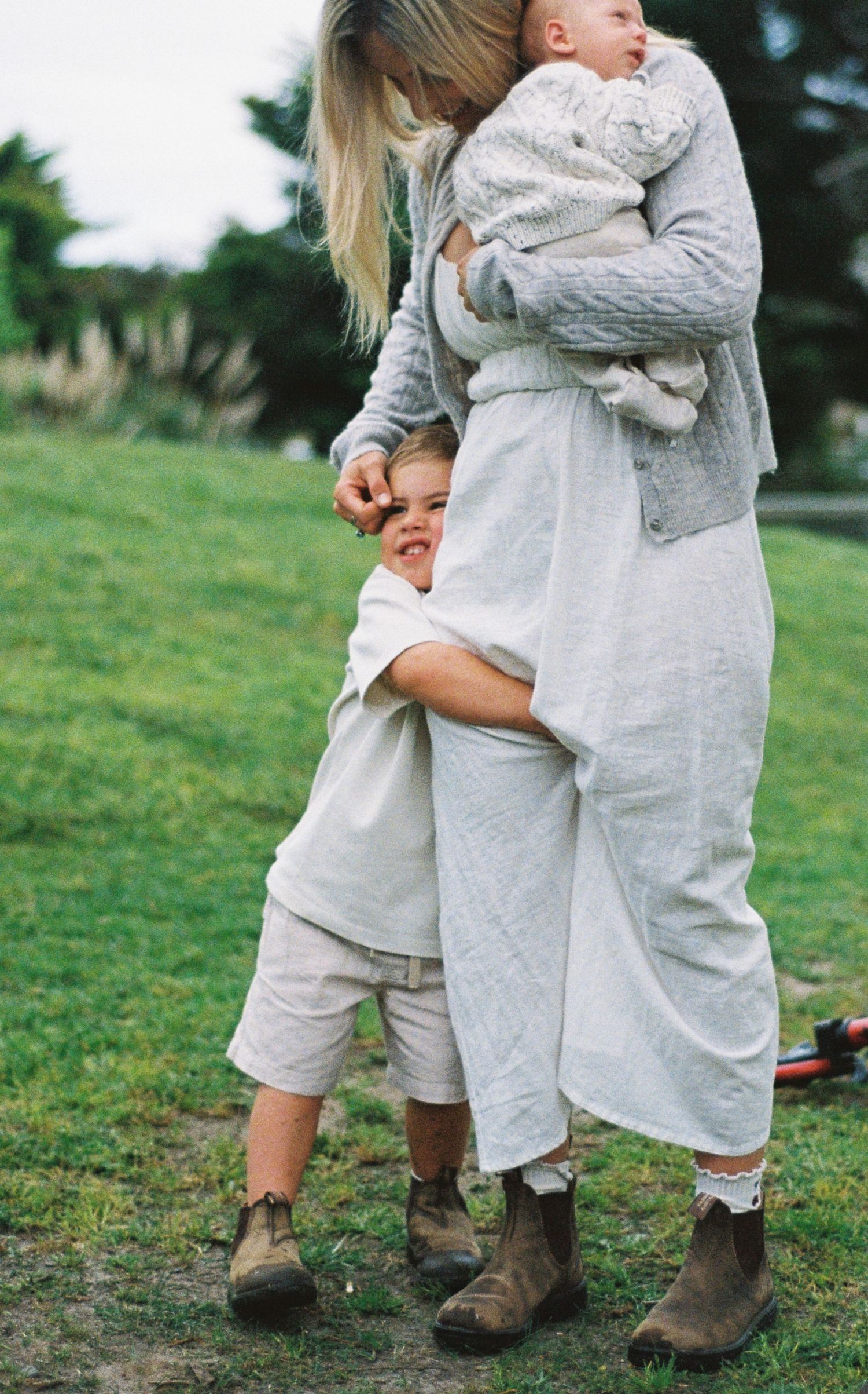 A blonde woman in linen clothing holds a baby while a young child hugs her legs outdoors on green grass.