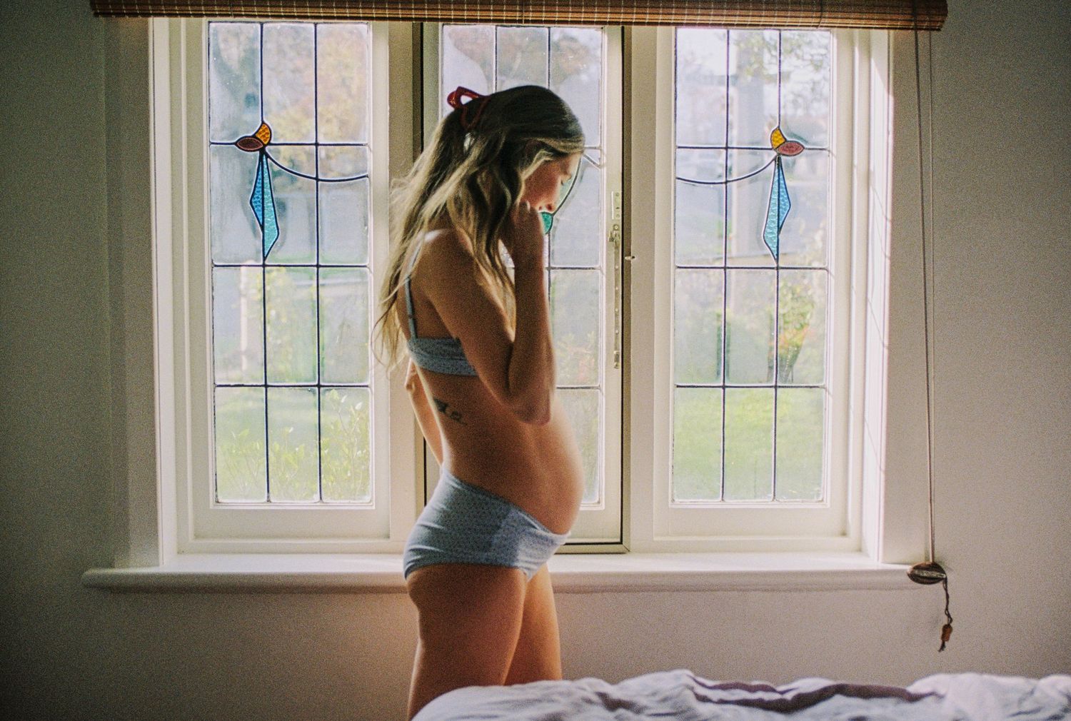 Pregnant woman in blue lingerie standing by stained glass window in bedroom, side profile showing baby bump.