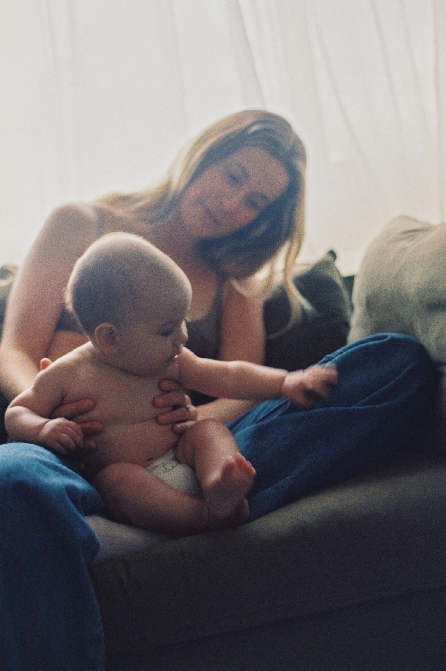 A blonde mother tenderly holds her baby in a diaper while sitting on a couch near a bright window.