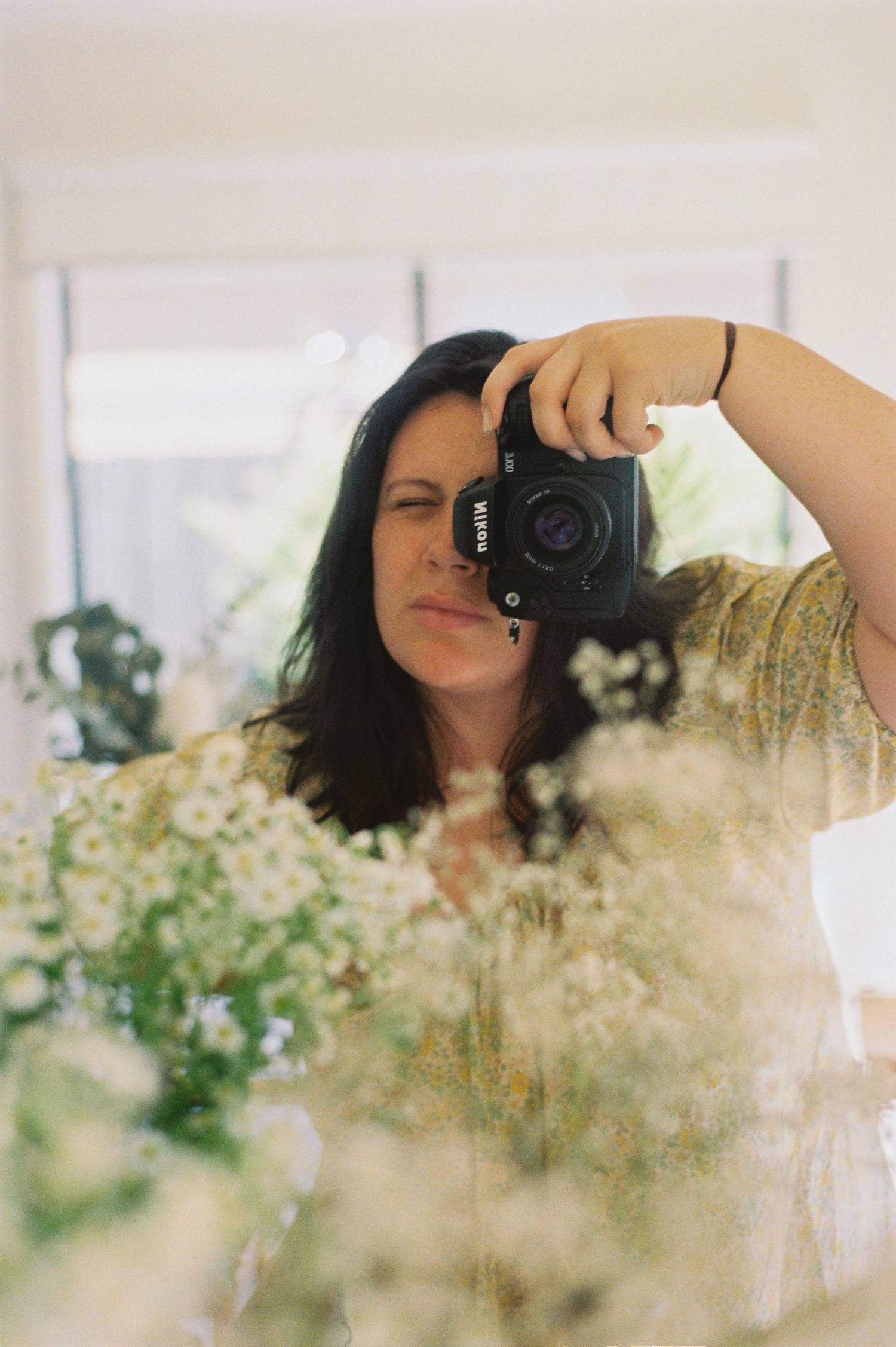 A photographer takes self portraits through a mirror with delicate white flowers in the foreground creating a dreamy effect.