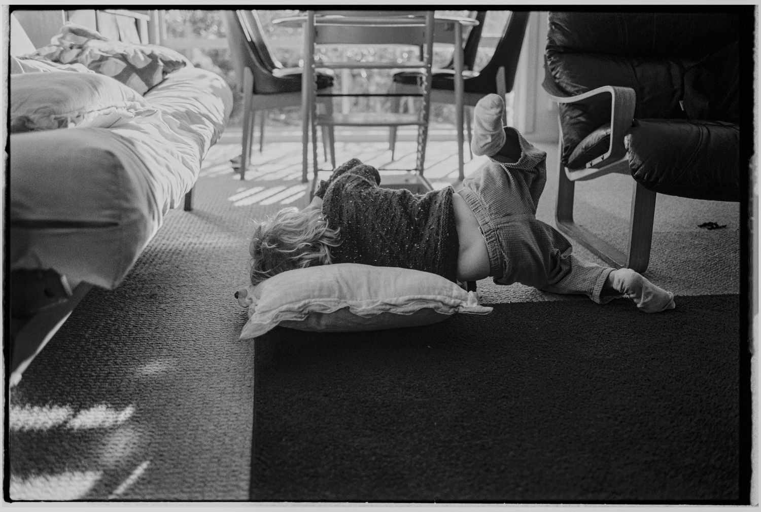 Black and white photo of a person lying on floor pillows in a sunlit room with beds and chairs nearby.