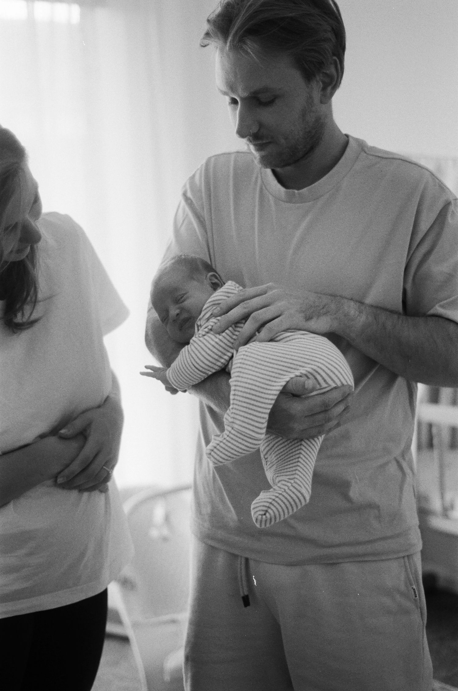 Black and white photo of a man tenderly holding a newborn baby while a woman stands nearby indoors.