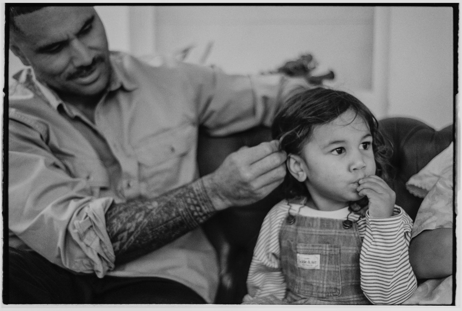 Tattooed father tenderly fixes young daughter's hair while she sits eating in black and white photo.