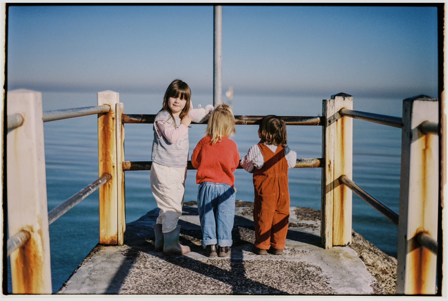 Three children stand on a seaside pier gazing at calm blue ocean waters on a sunny day.