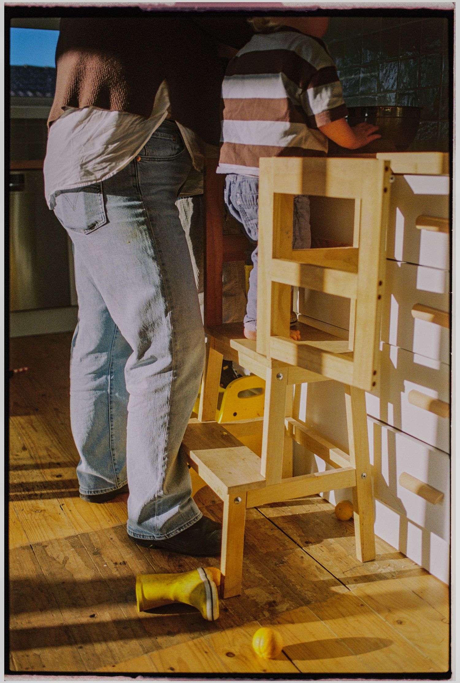 Adult and child standing beside a wooden learning tower on hardwood floors, with yellow toy visible nearby.