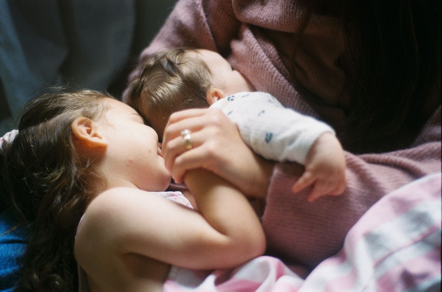 A mother breastfeeding a newborn baby while an older toddler cuddles closely beside them.