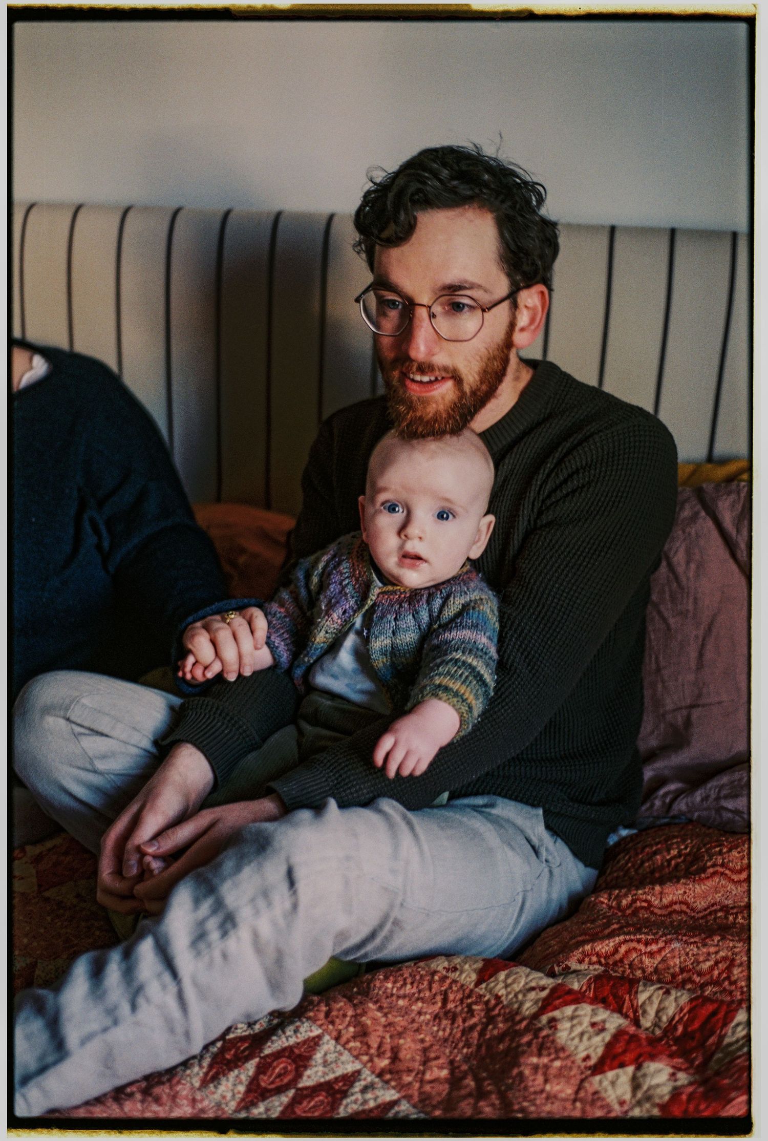 Bearded man with glasses sits cross-legged on a bed holding a baby in a colorful knit sweater.