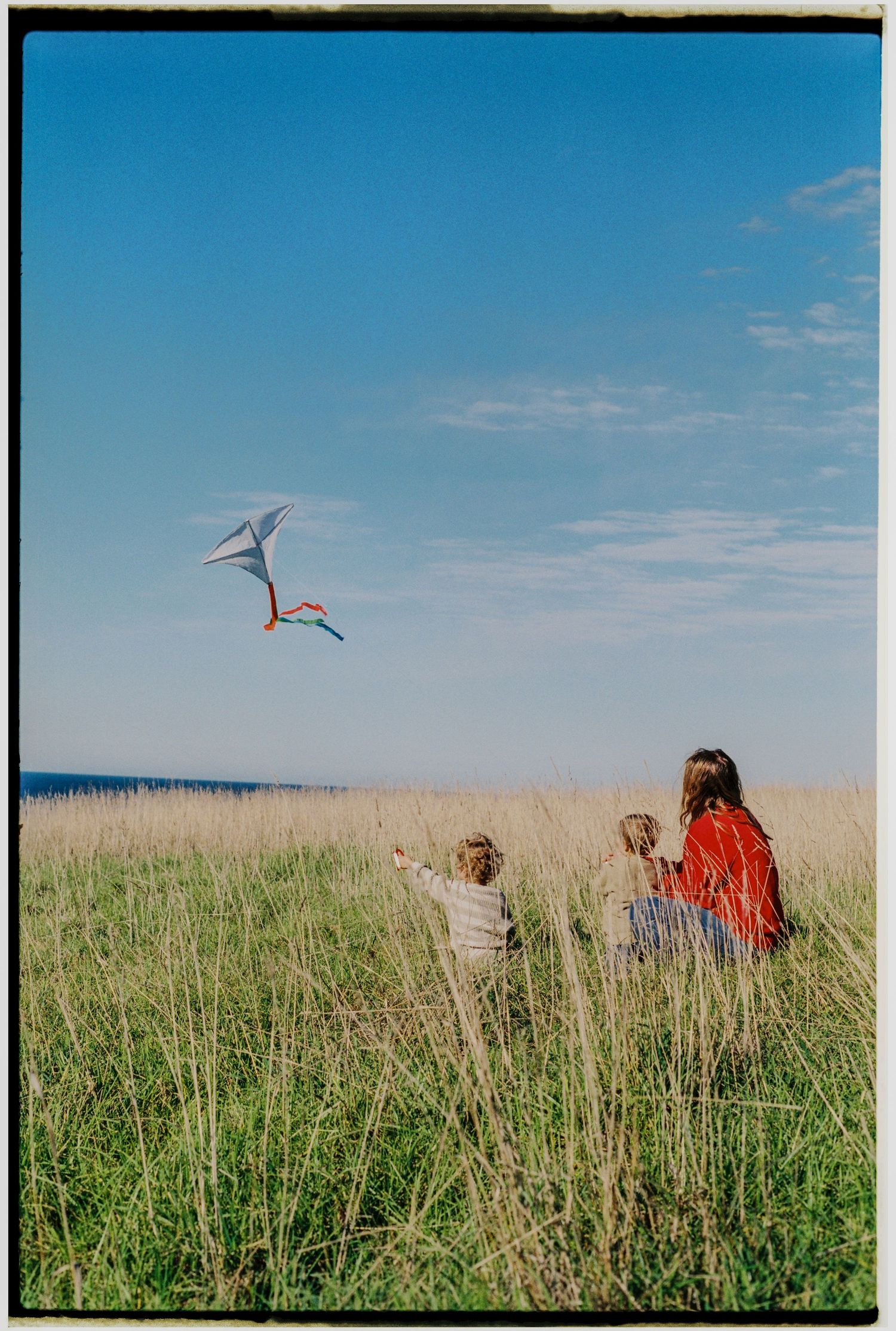 A mother and two children sit in tall grass flying a colorful kite against a bright blue sky.