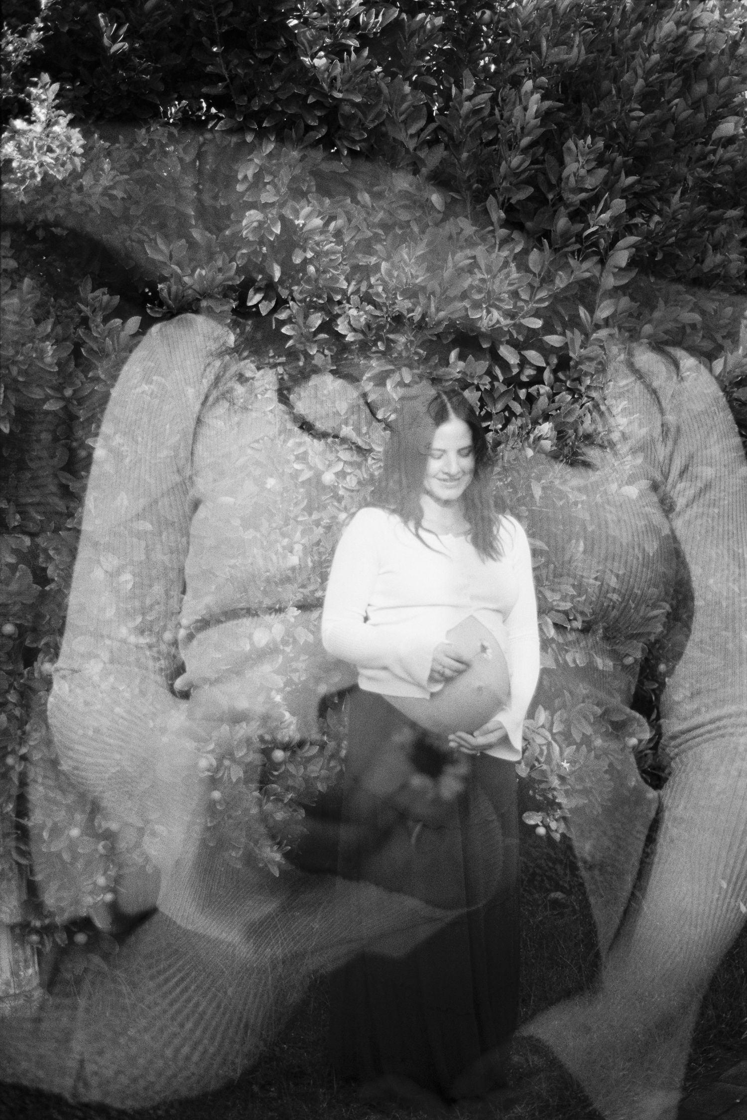 Black and white double exposure photo of a pregnant woman cradling her belly, surrounded by lush foliage.
