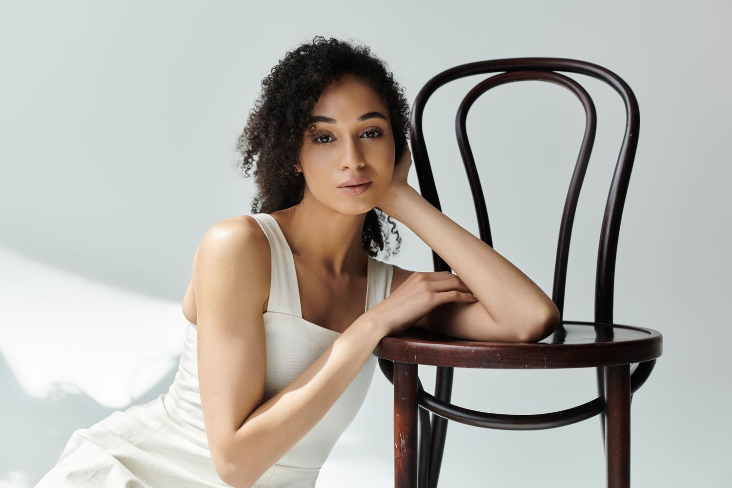 Person in white outfit posing elegantly with wooden chair against light background.