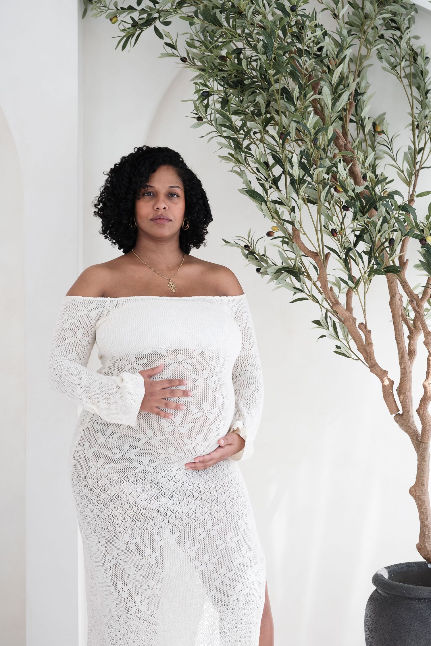 Elegant maternity photoshoot featuring a white off-shoulder lace gown against a backdrop of artificial olive trees.