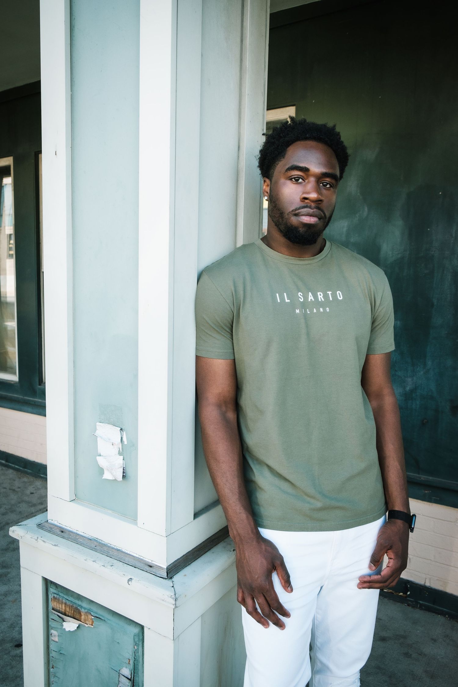 A man wearing an olive green t-shirt and white pants poses against a wall in three similar photos.