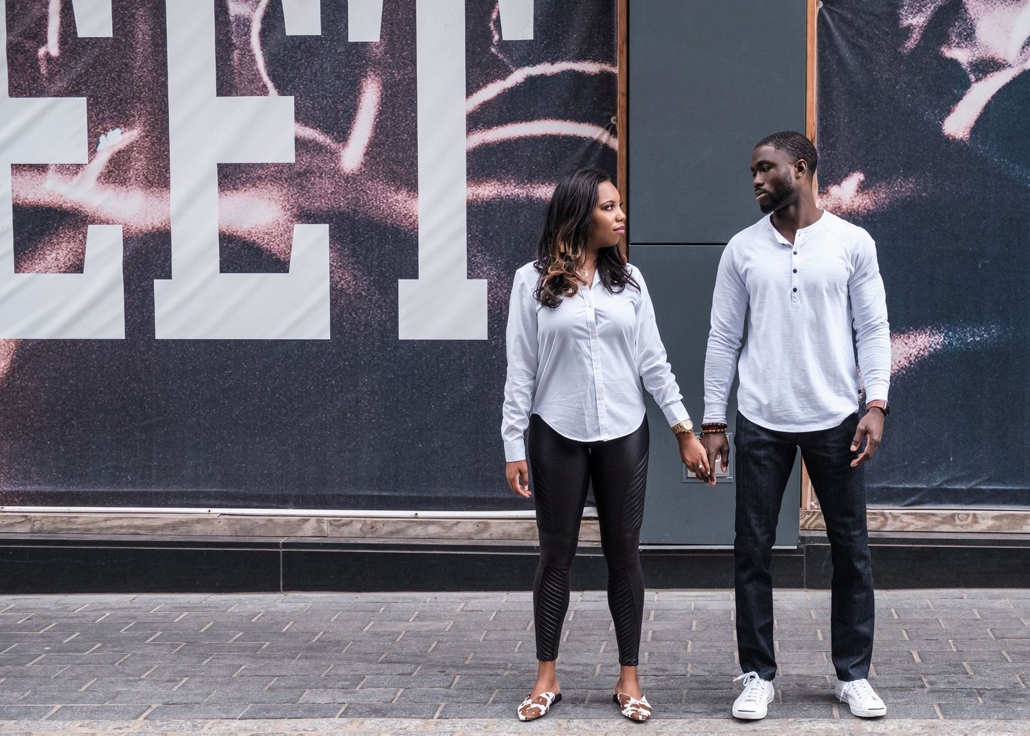 A stylish couple in coordinated white outfits poses against an urban wall with artistic graphics.