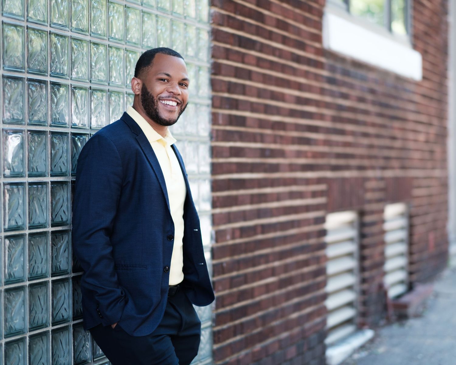 A professional wearing a navy suit and cream tie smiles while leaning against a brick wall outdoors.