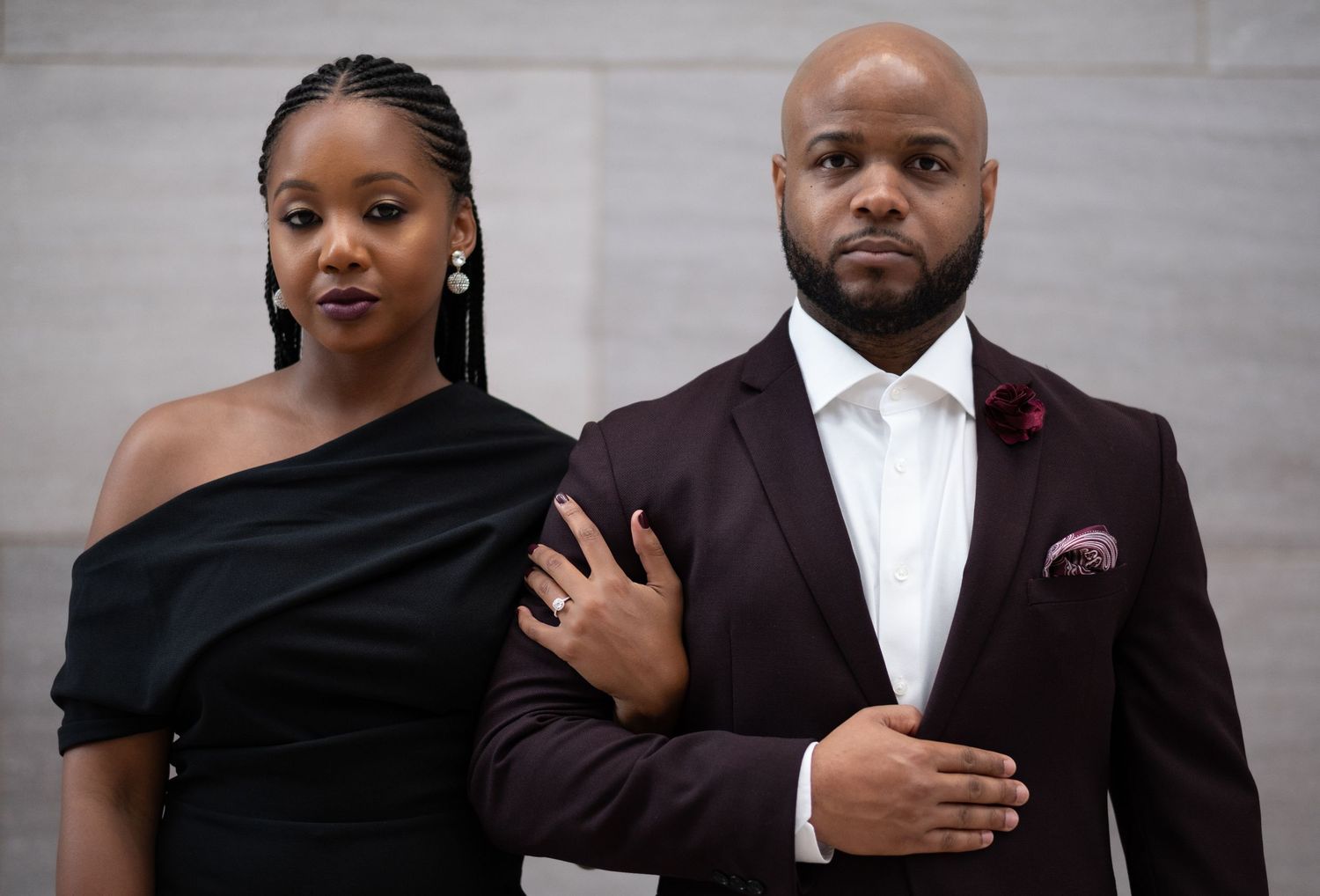 Couple in contrasting black and burgundy formal wear posing against a light gray wall.
