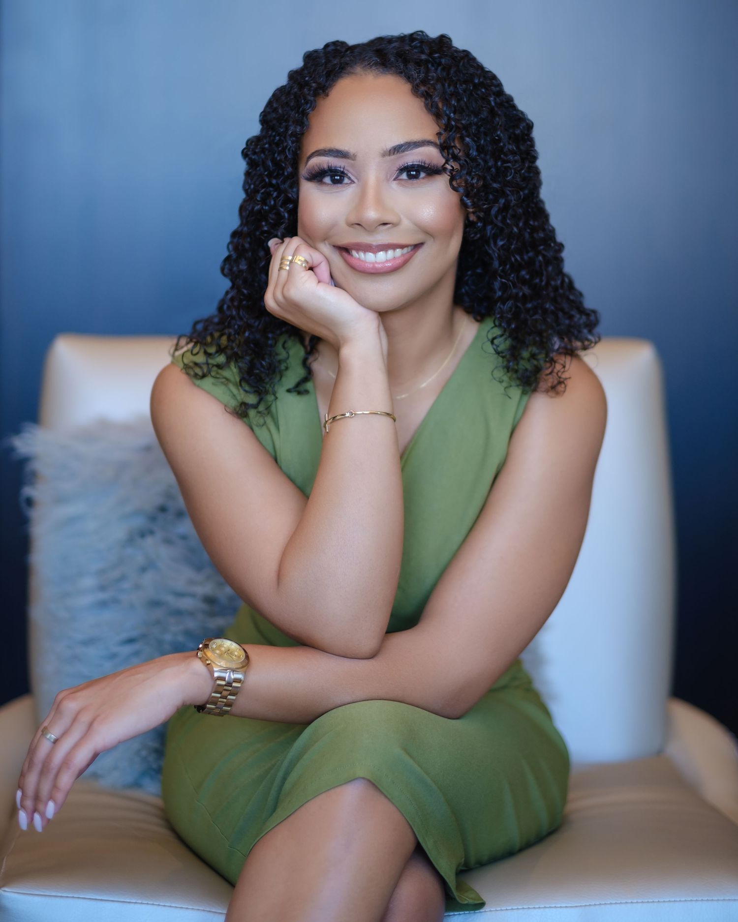 A person in a green dress poses confidently on a beige couch in a professional photoshoot setting.
