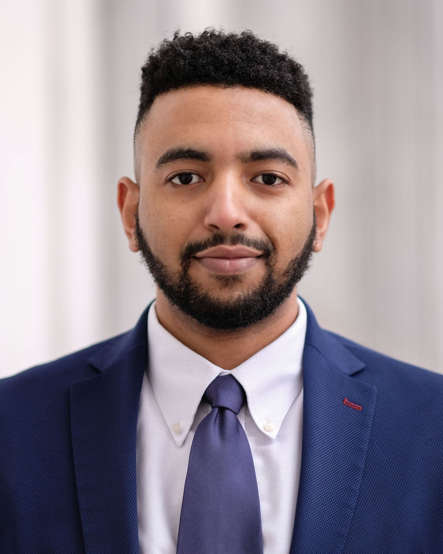 Professional headshot of person in navy suit and tie against light background.