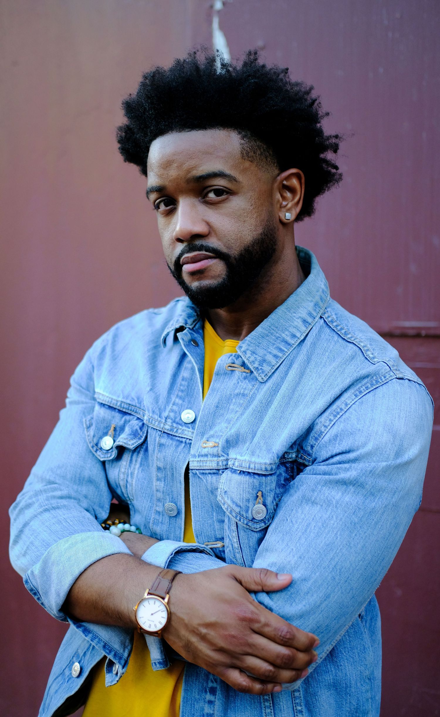 A person wearing a light denim jacket and yellow shirt poses against a purple wall in a photo series.