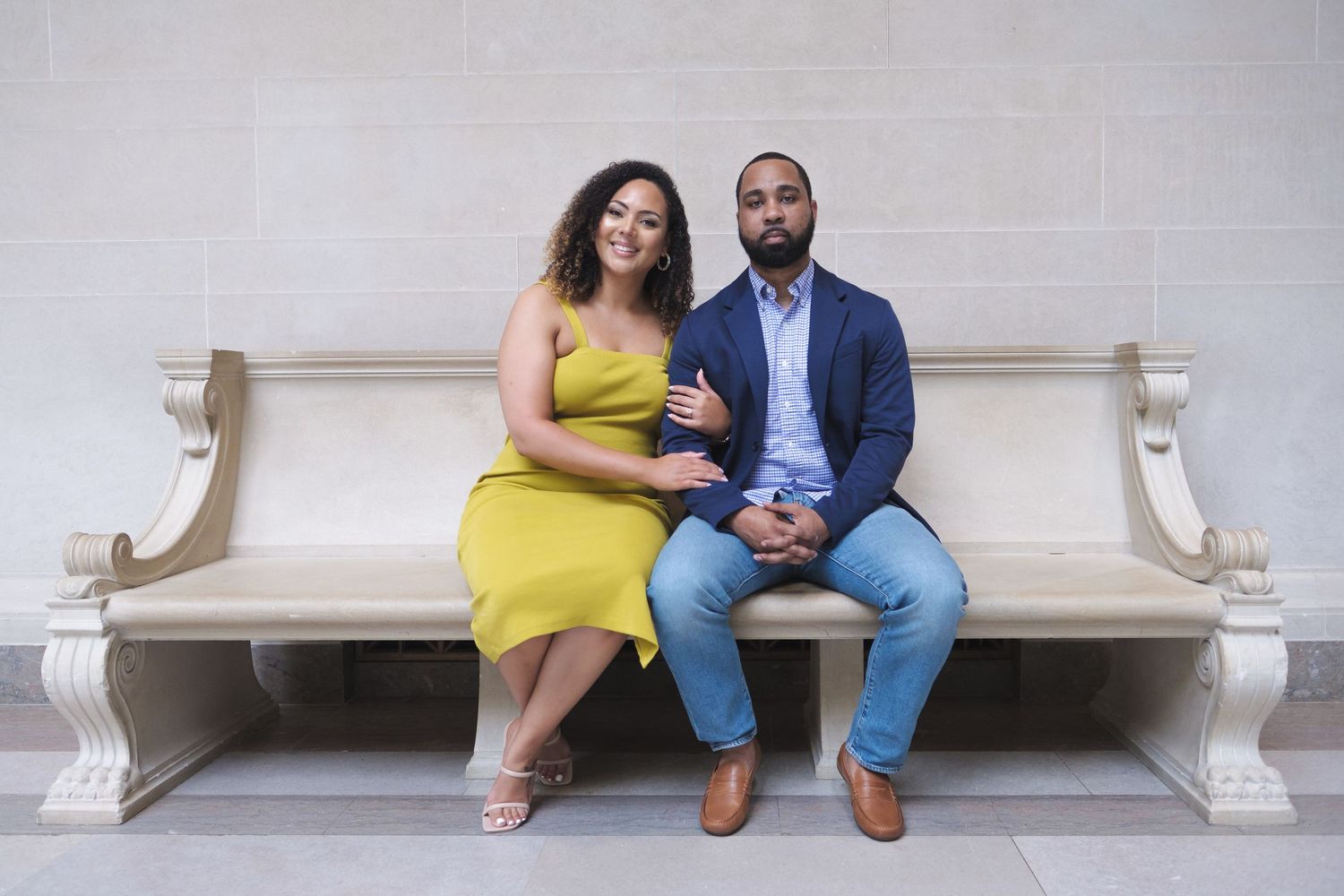 Couple in yellow dress and blue suit sitting together on a white stone bench.